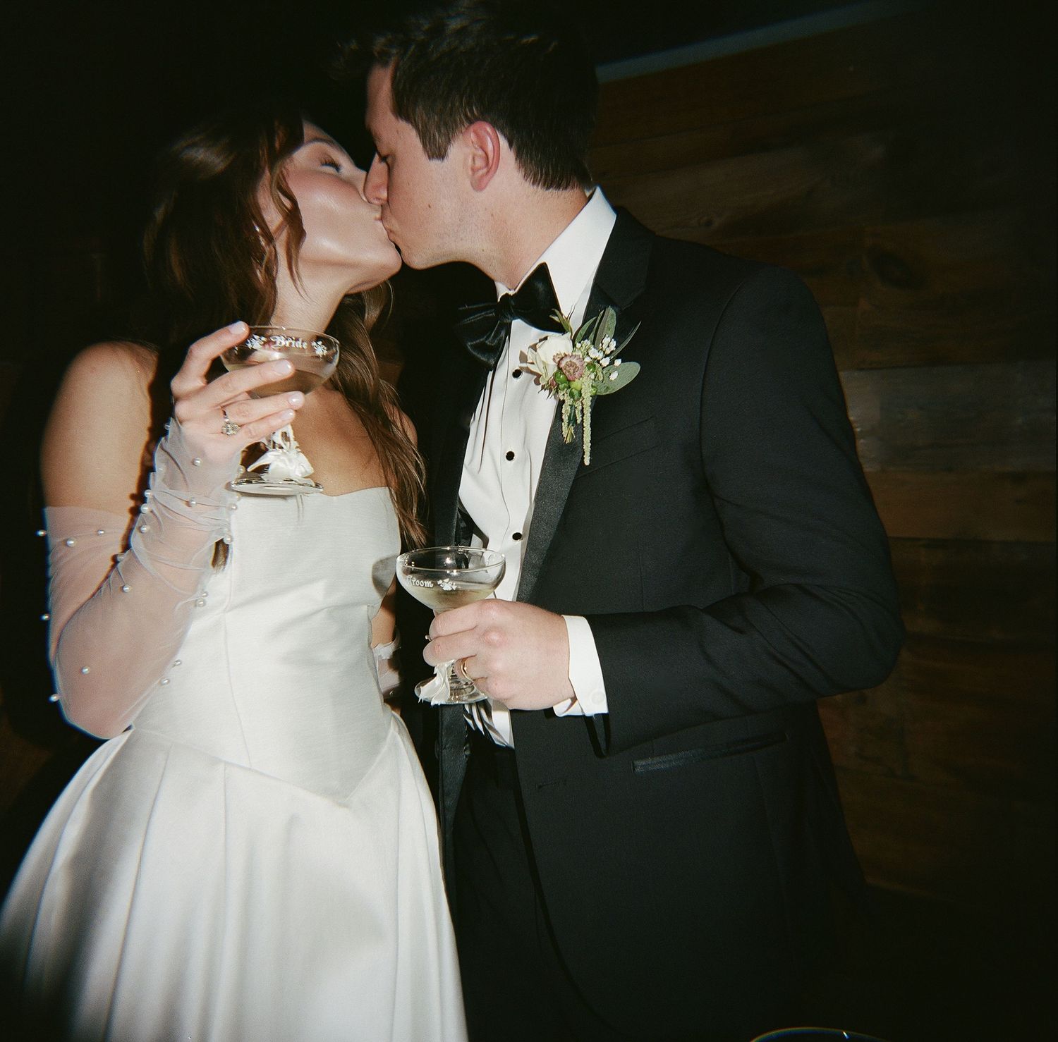 A bride and groom share a romantic kiss while holding champagne glasses at their wedding reception.