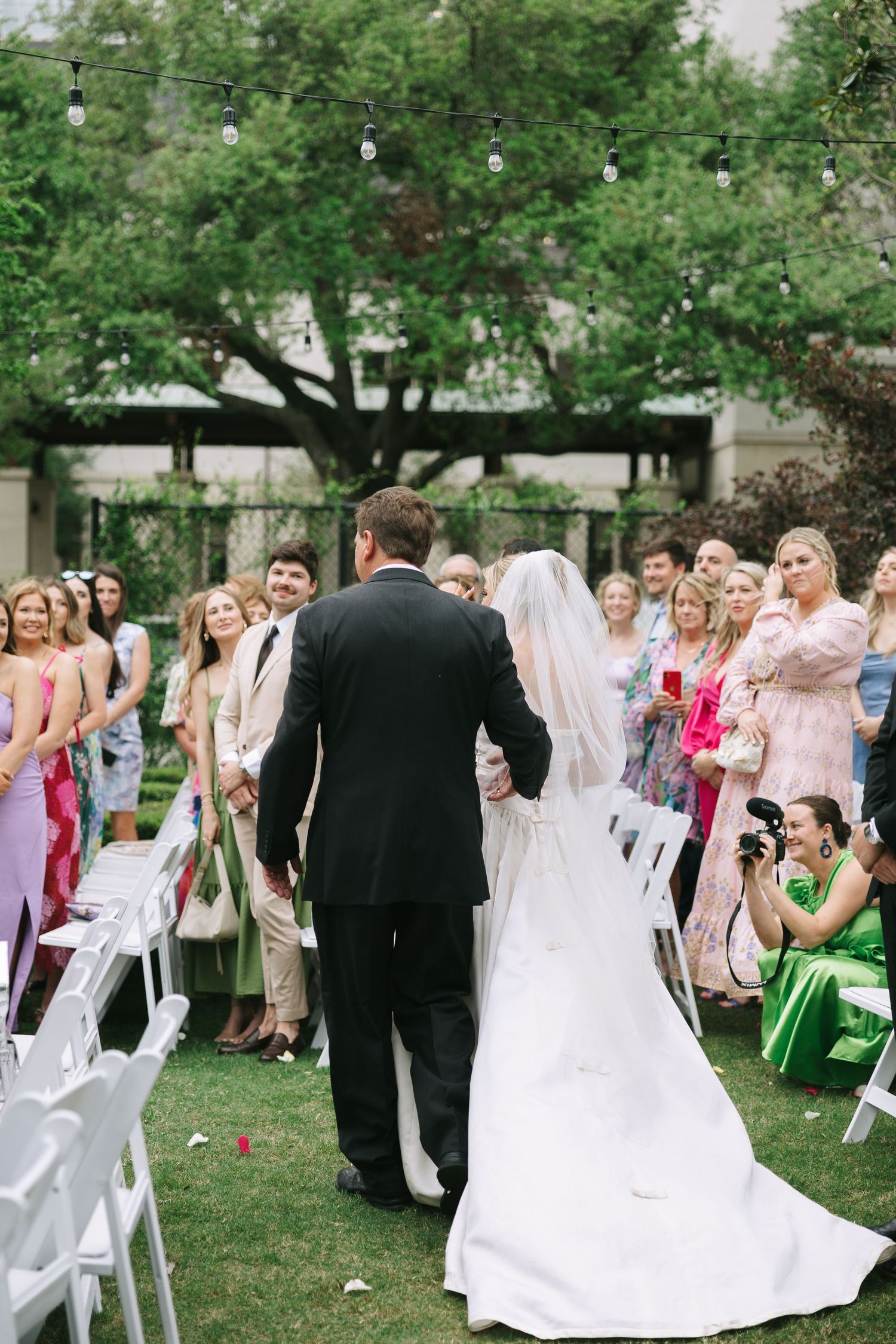 Wedding ceremony processional with guests seated on white chairs on garden lawn.