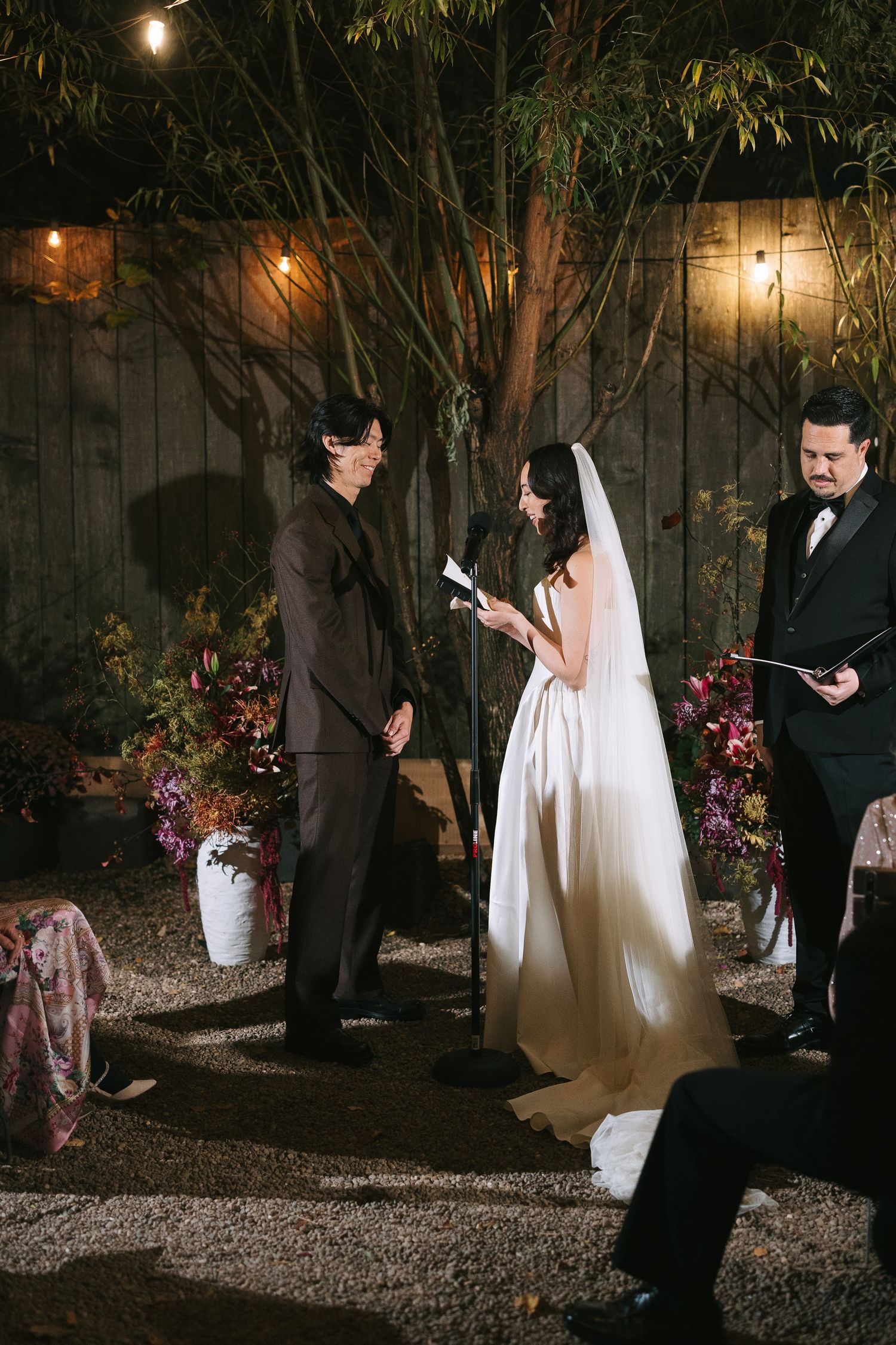A wedding ceremony takes place in a garden setting with string lights and floral decorations.