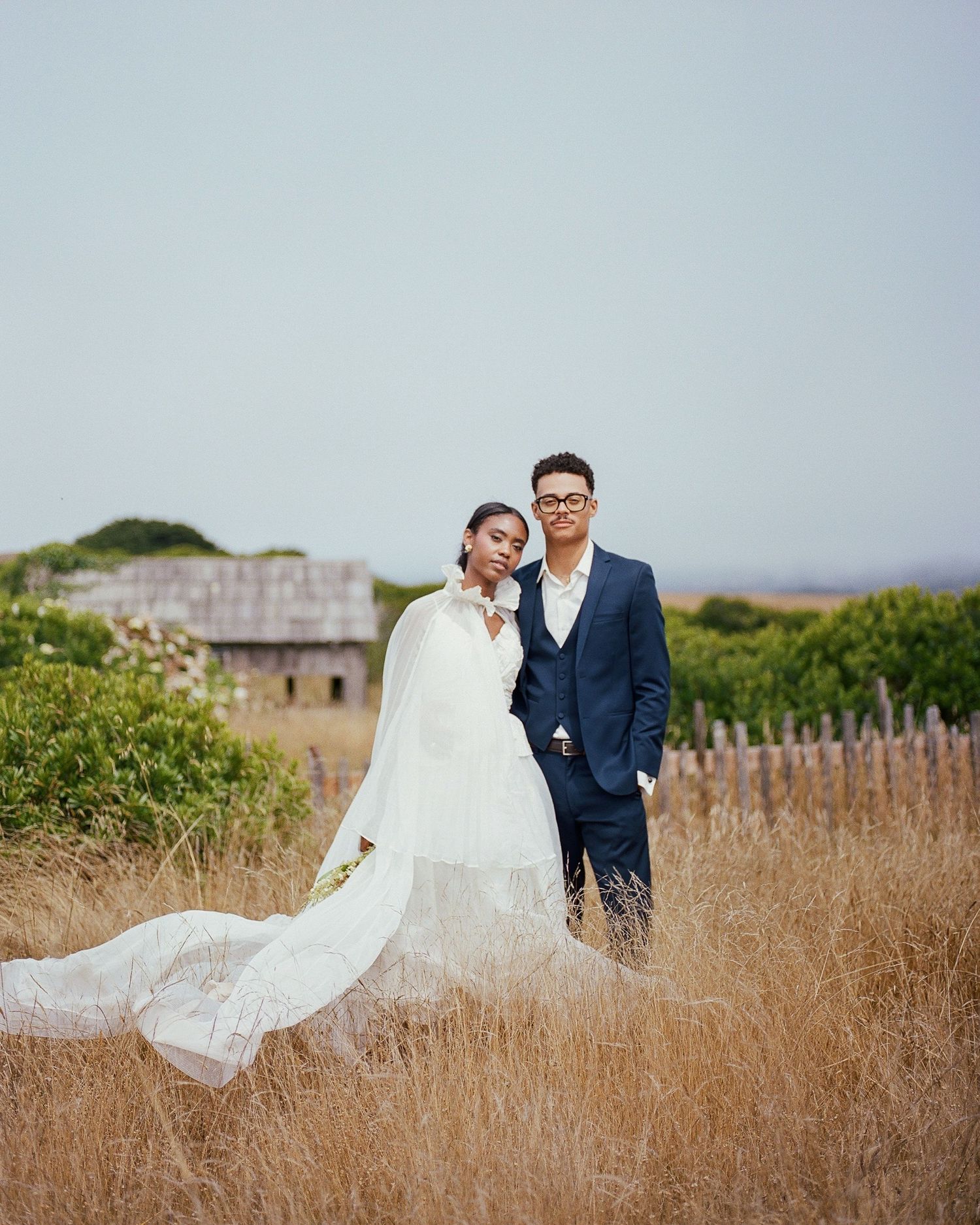 A bride in a white cape gown and groom in a navy suit pose romantically in a golden field near a rustic barn.
