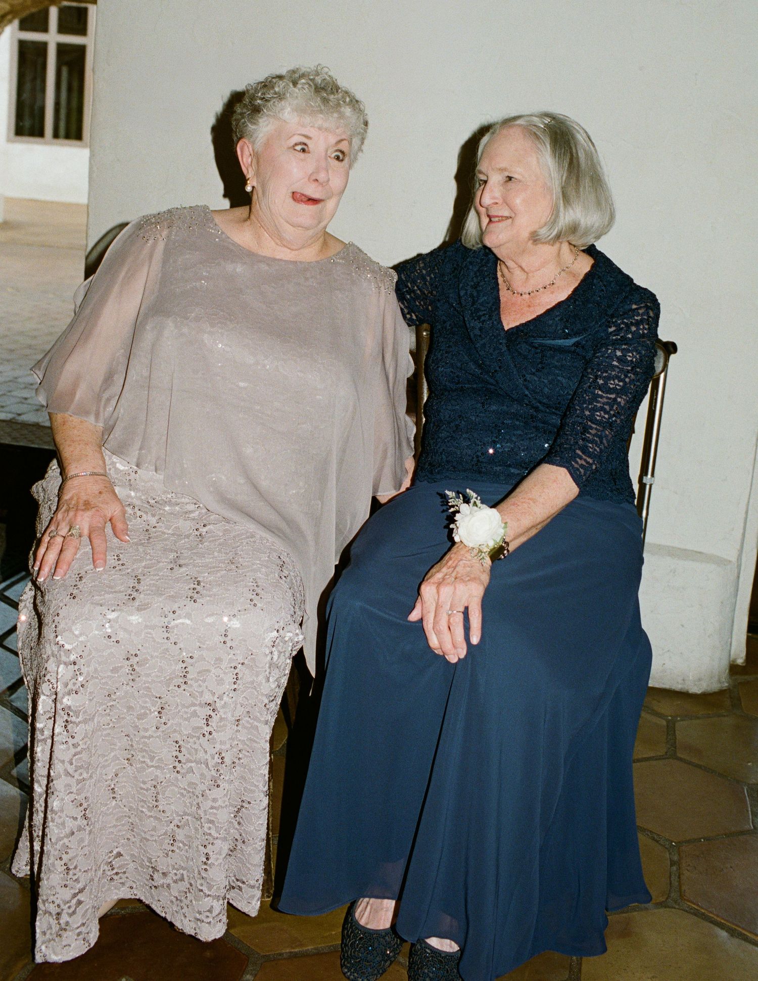 Two elderly women in formal attire sitting together, one in beige lace dress and one in navy blue gown with corsage.
