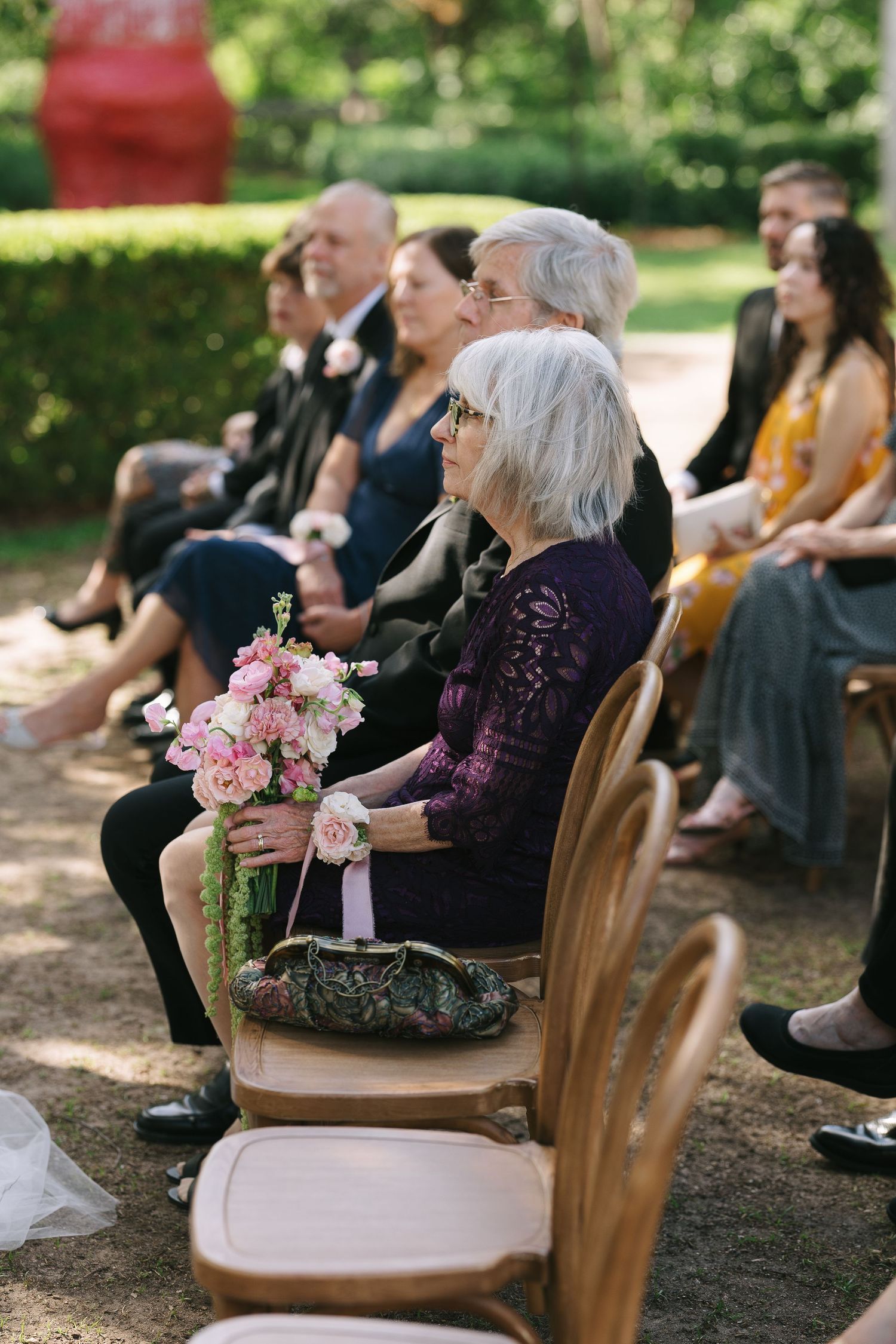 Guests seated in wooden chairs at outdoor ceremony with pink floral arrangements.