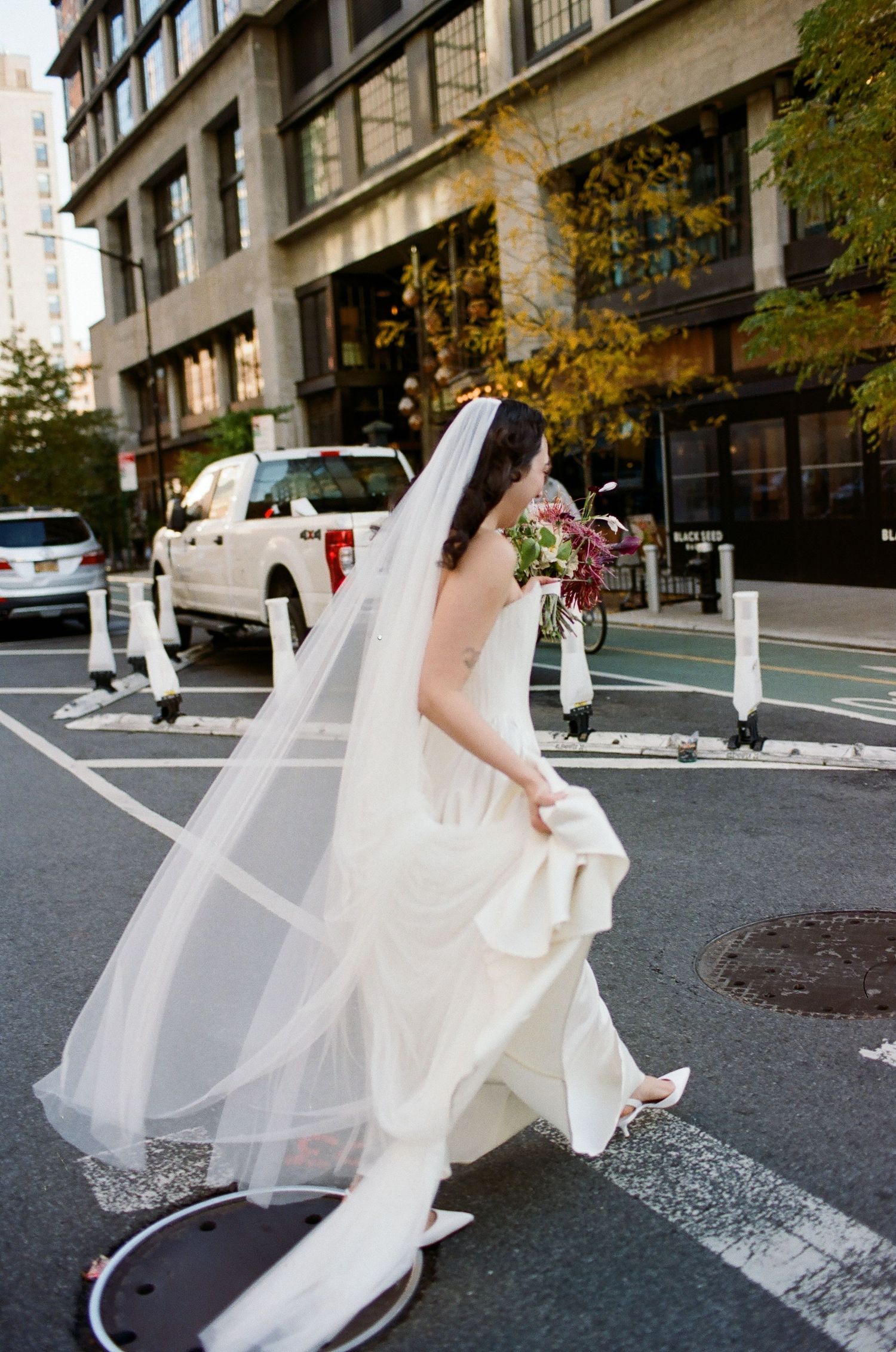 A flowing white wedding dress and veil billow in the wind on a city street with autumn trees in the background.