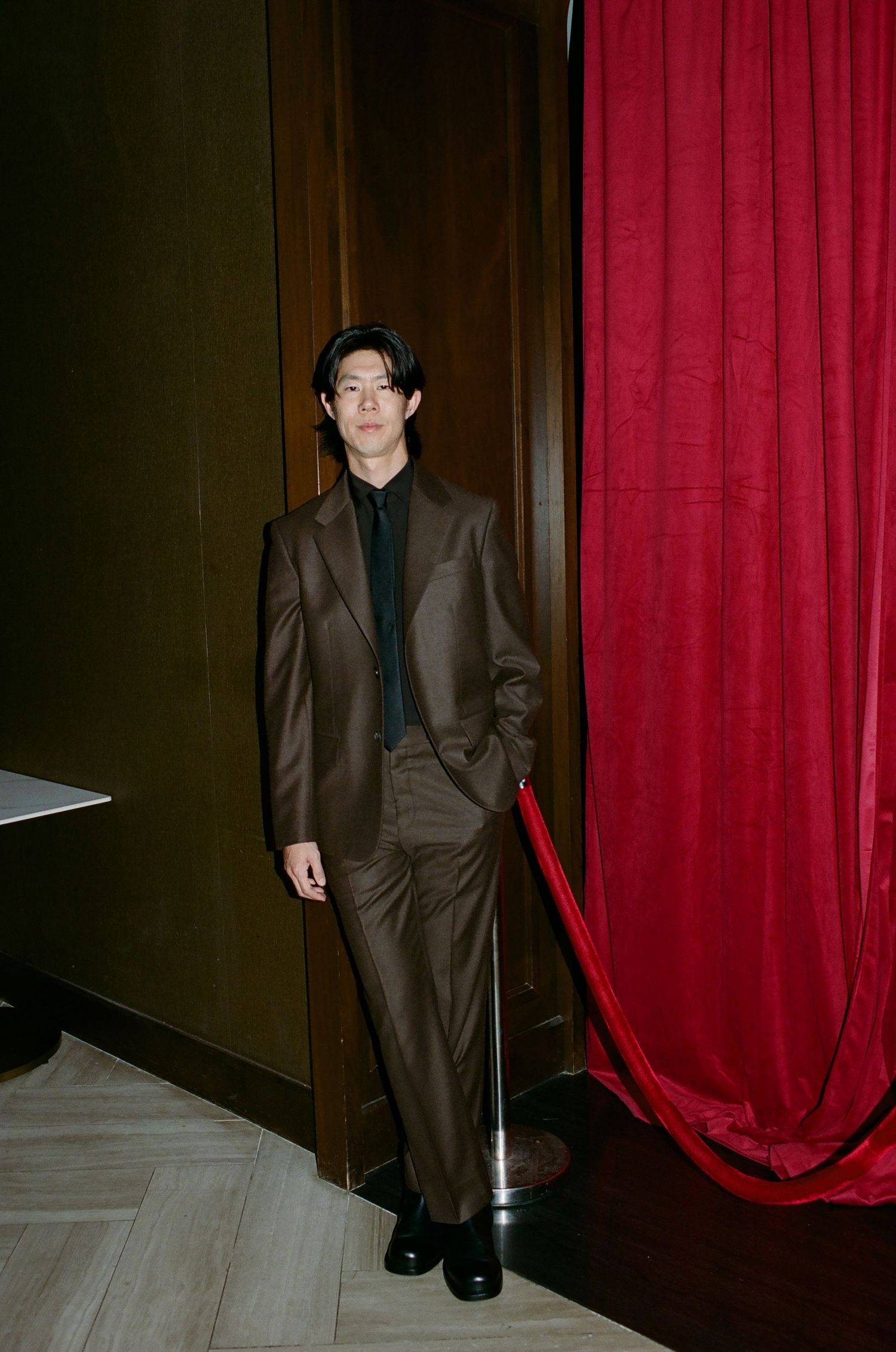 A person in a brown suit stands elegantly against a backdrop of wooden walls and red velvet curtains.