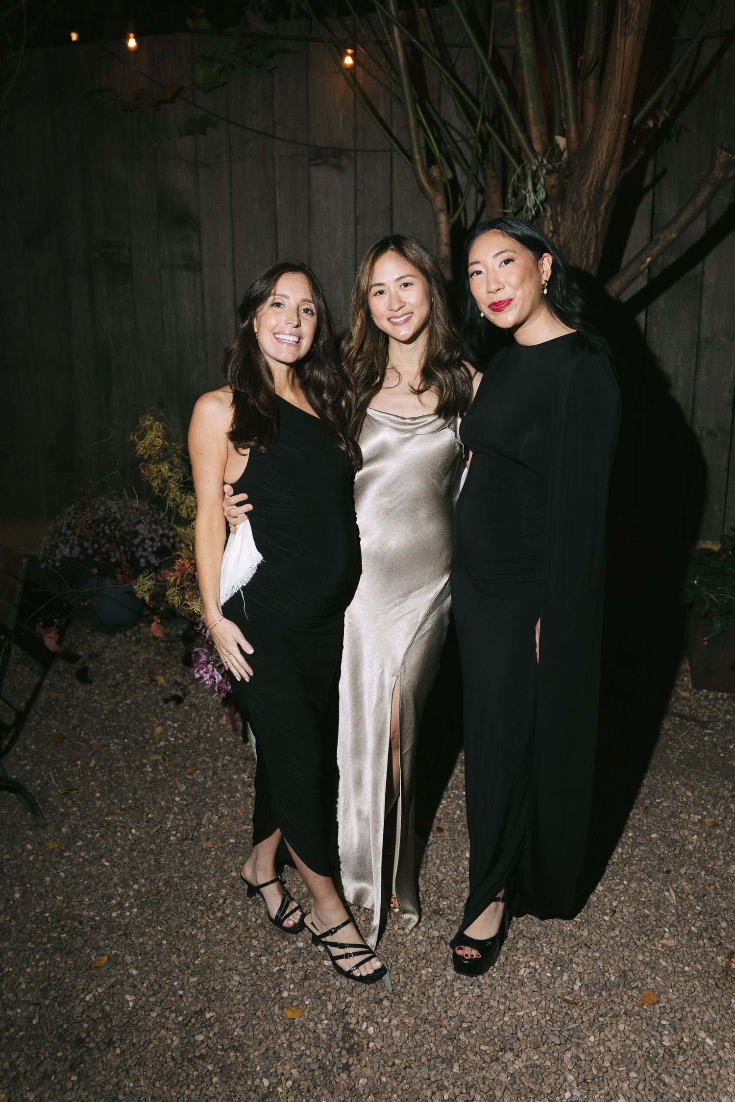 Three women pose together at an evening event wearing elegant dresses in black, champagne silk, and black.