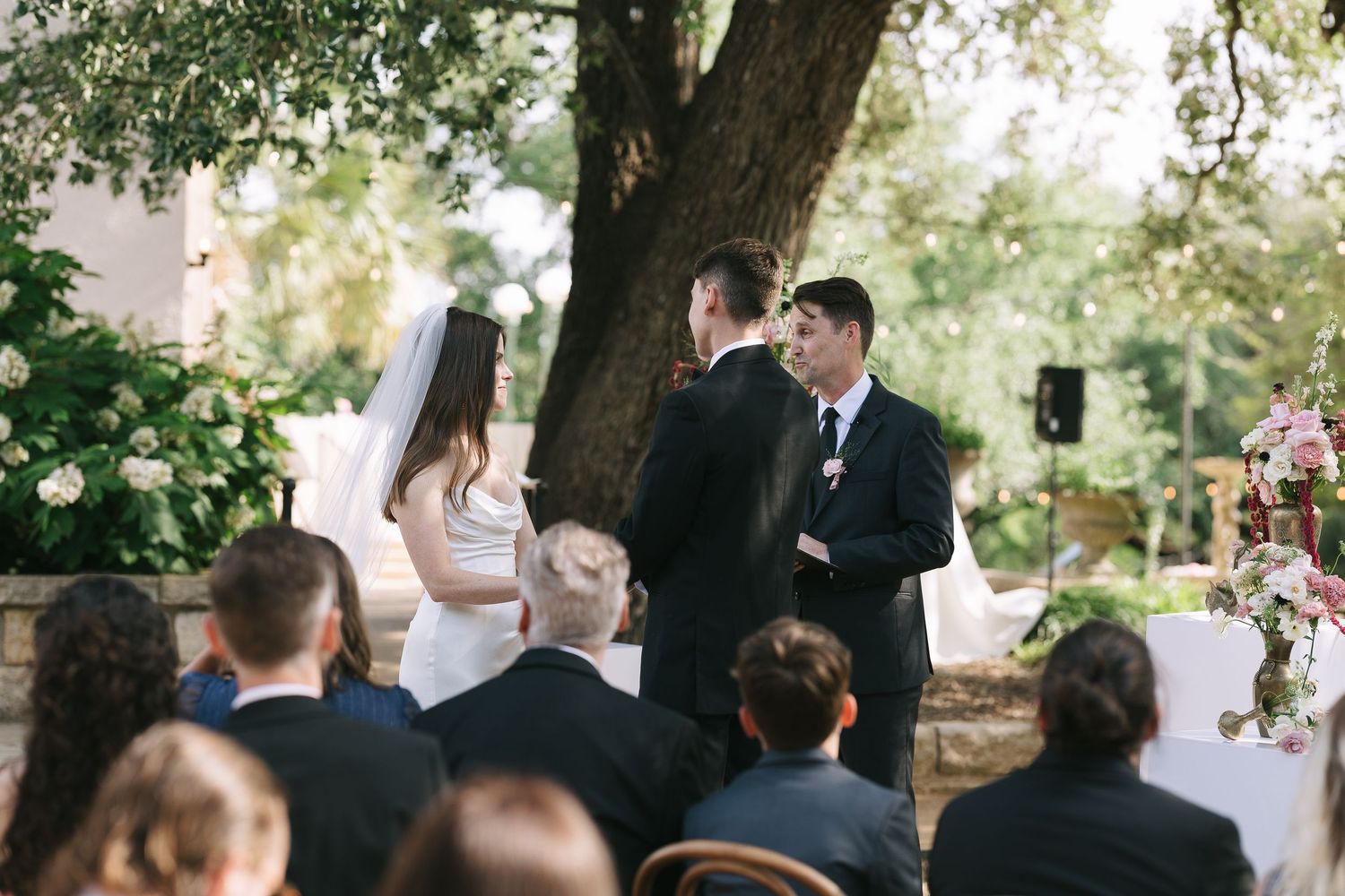 Outdoor wedding ceremony setup with guests seated under green tree canopy.
