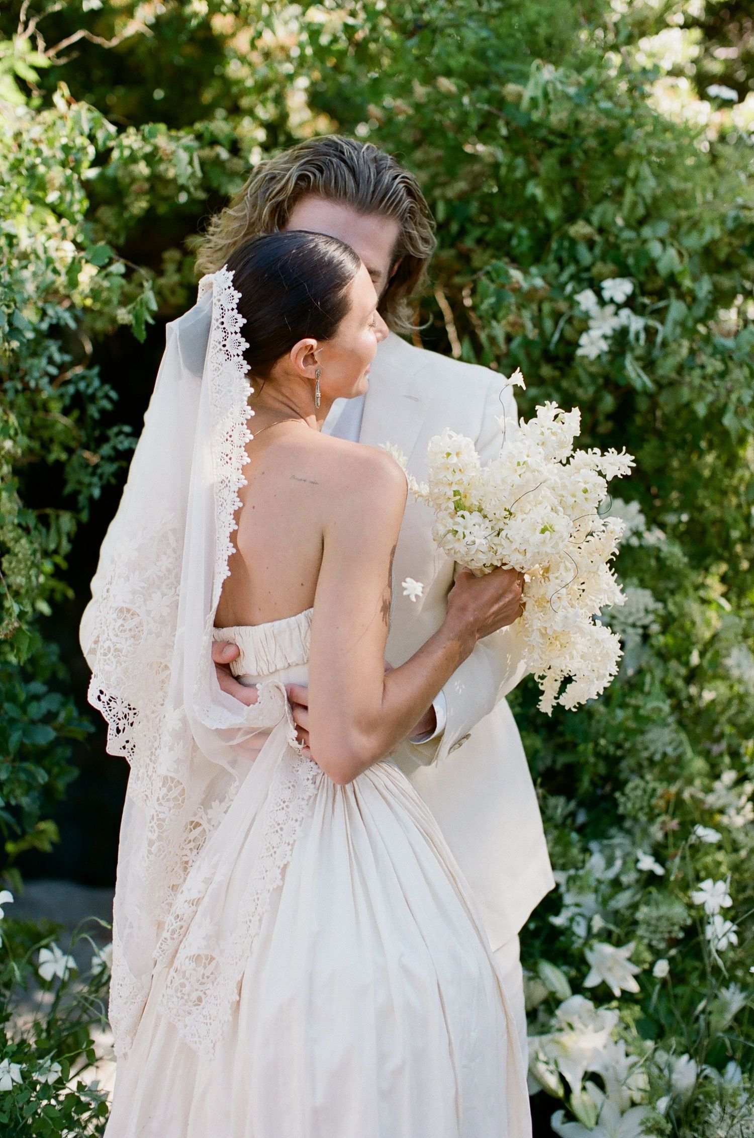Bride and groom embrace outdoors, her lace veil and white floral bouquet complementing lush green garden backdrop.