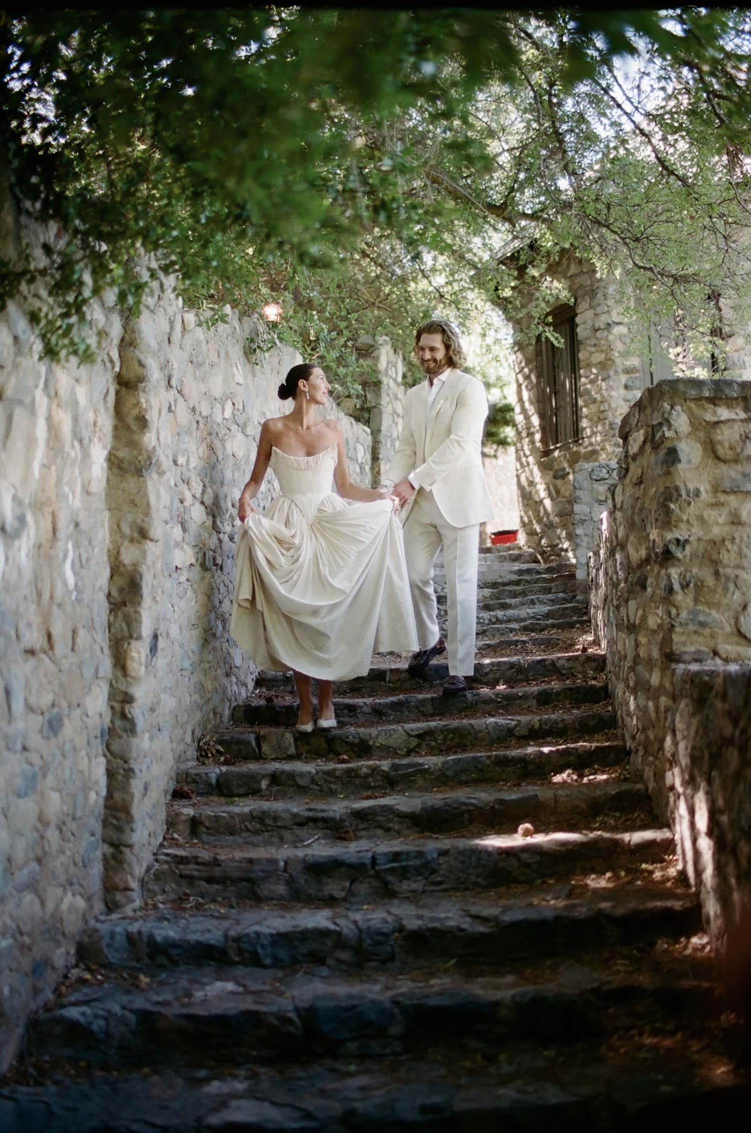 Bride and groom walking up stone steps in a garden, bride lifting her white gown, groom in white suit.