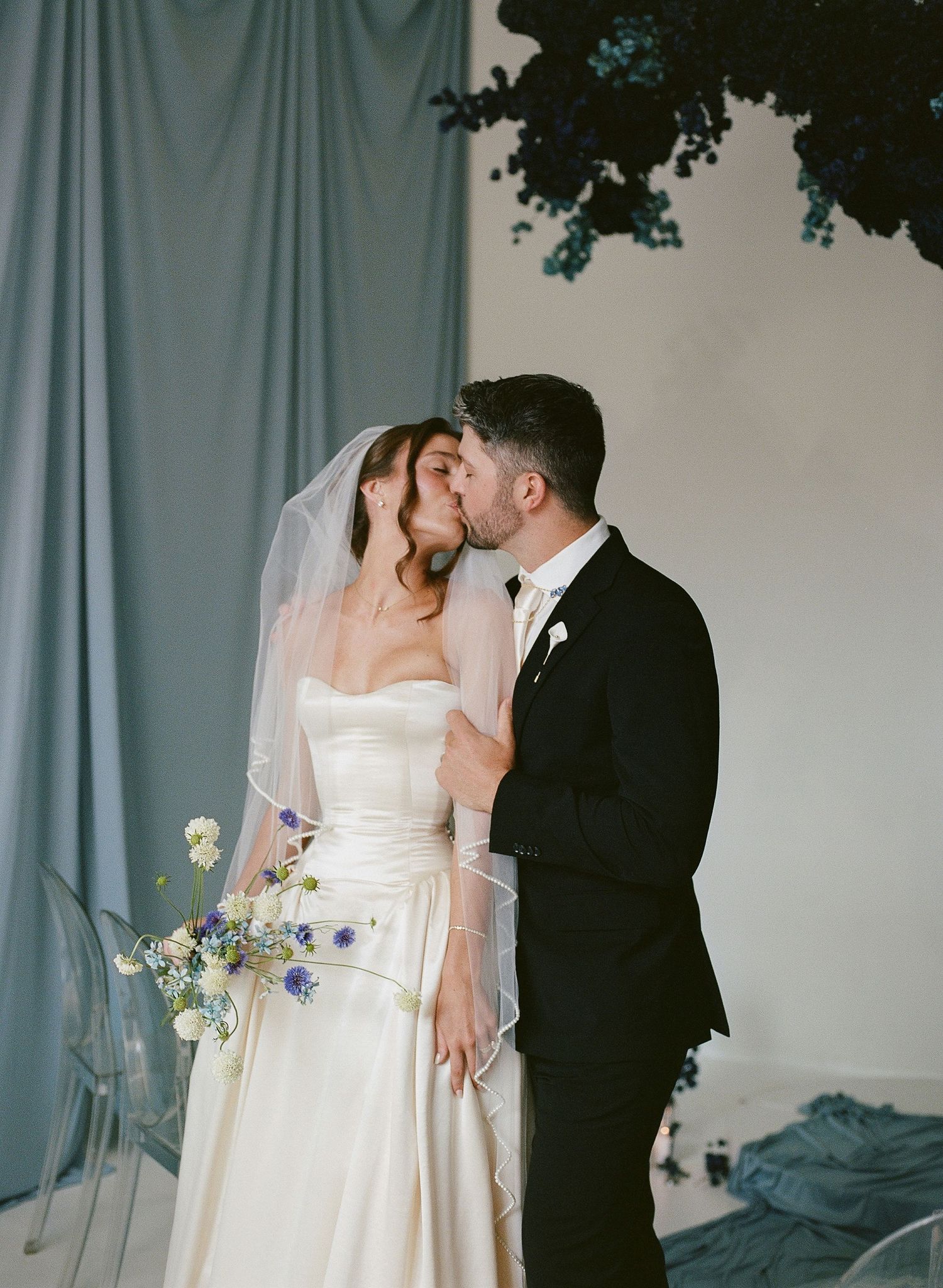 Bride and groom share a kiss at their wedding, bride wearing a white strapless gown with veil holding a blue floral bouquet.