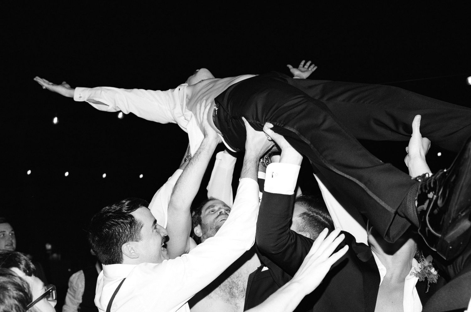 Black and white photo of wedding guests crowd surfing the groom at a lively reception celebration.