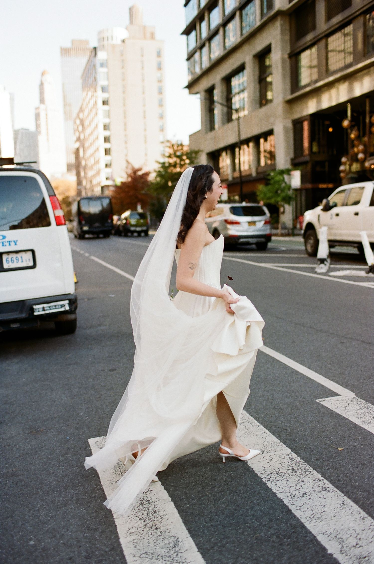 Bride in white gown and veil crossing a busy New York City street, holding her dress while cars and buildings surround her.