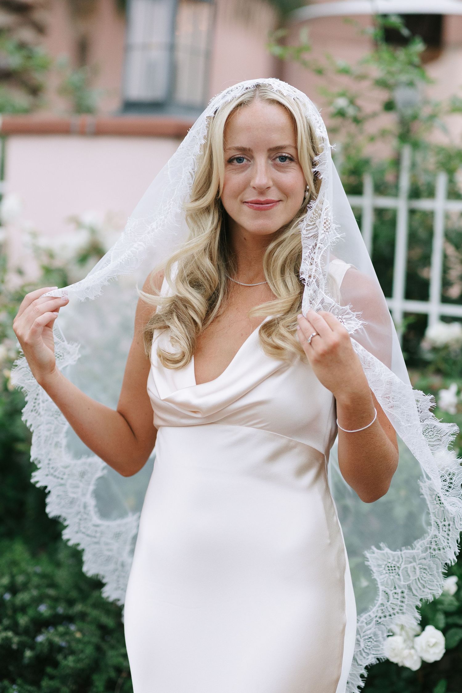 A joyful bride in white dress and lace veil poses against a pink wall and garden backdrop.