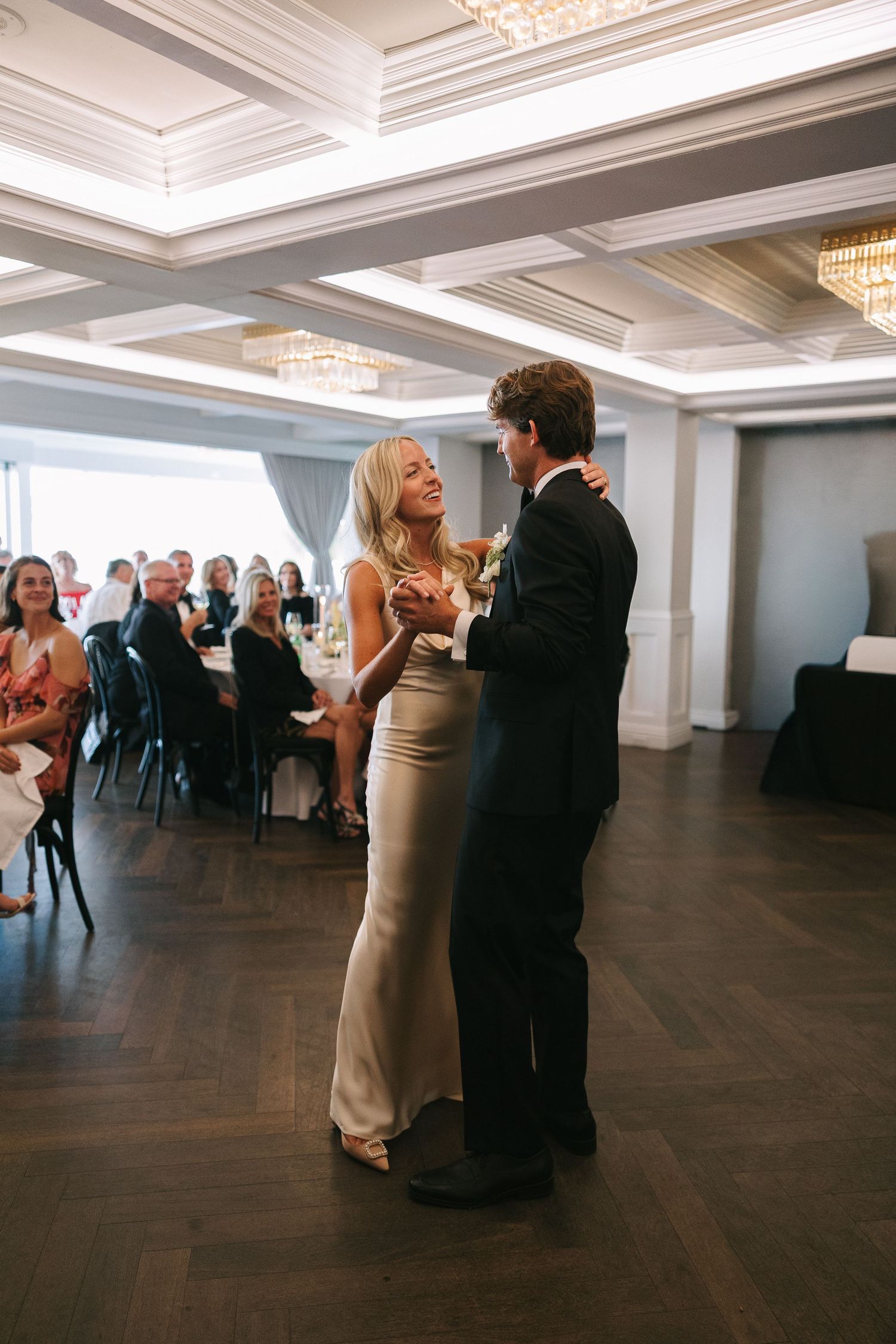 Couples dancing on dark hardwood floor under crystal chandeliers at formal event.