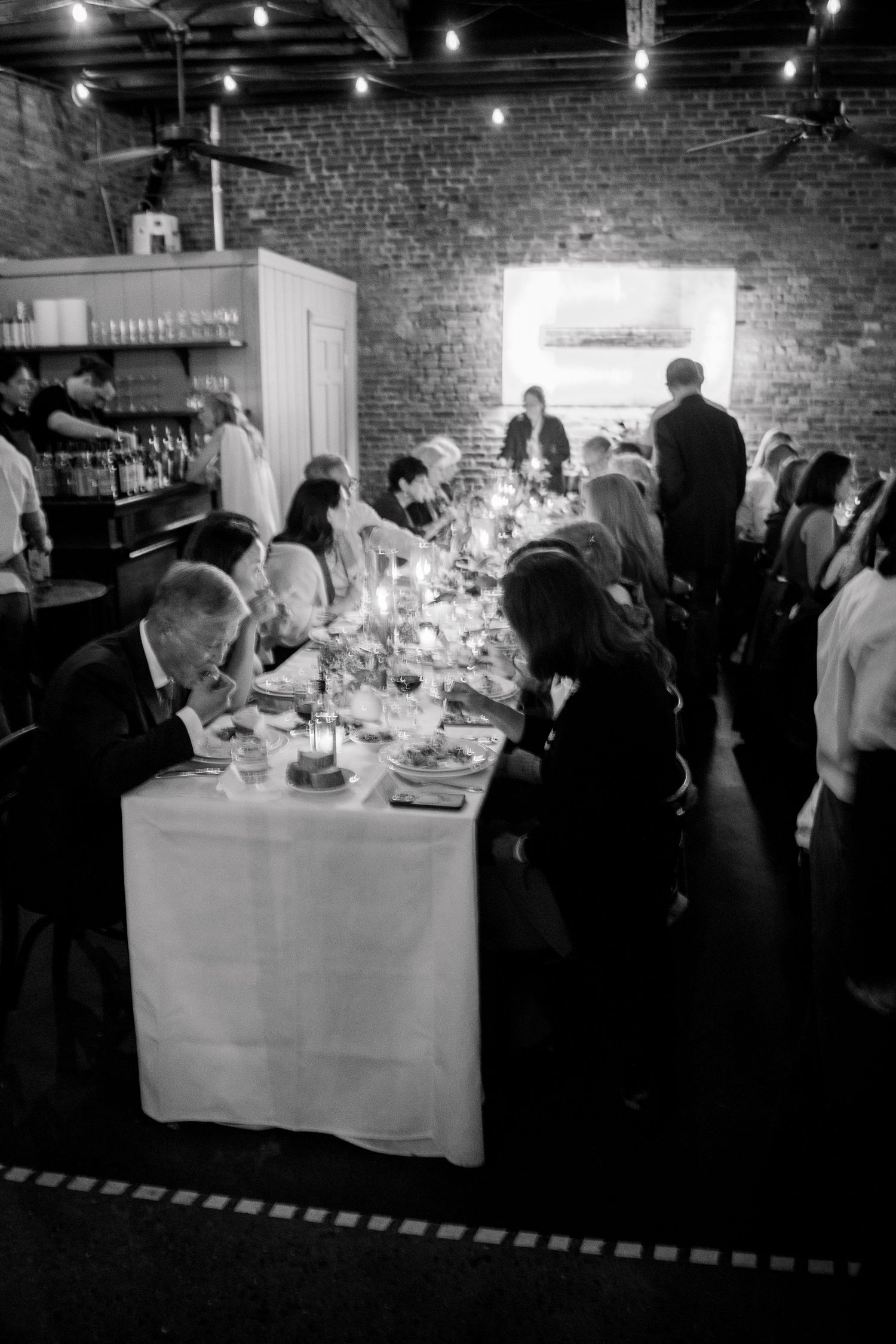 People gathered around formal dining tables in an industrial-style venue with exposed brick walls.