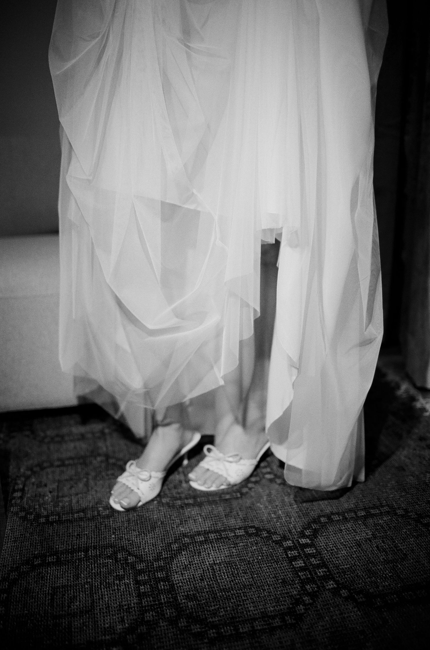 Black and white photo of a bride in a flowing white gown wearing delicate heeled shoes on a patterned carpet.