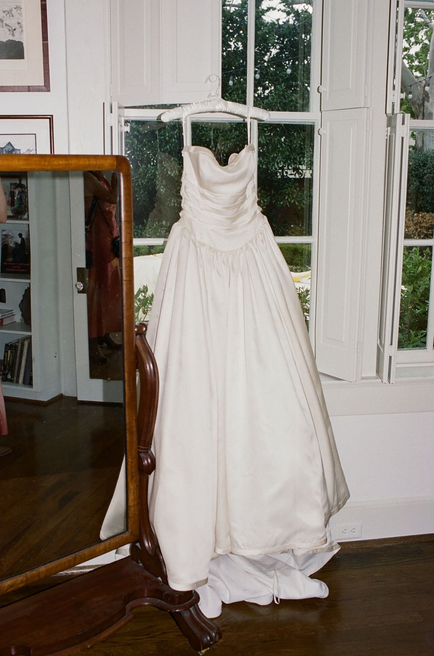 Elegant white wedding dress displayed in a bright room with large windows and hardwood floors.