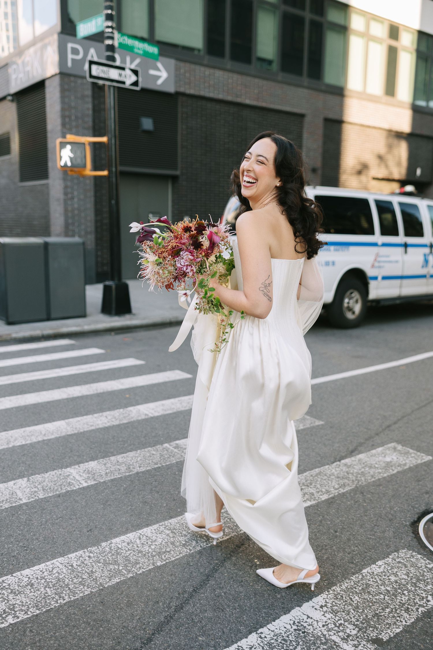 A bride dances across a crosswalk in an urban setting with her bouquet.