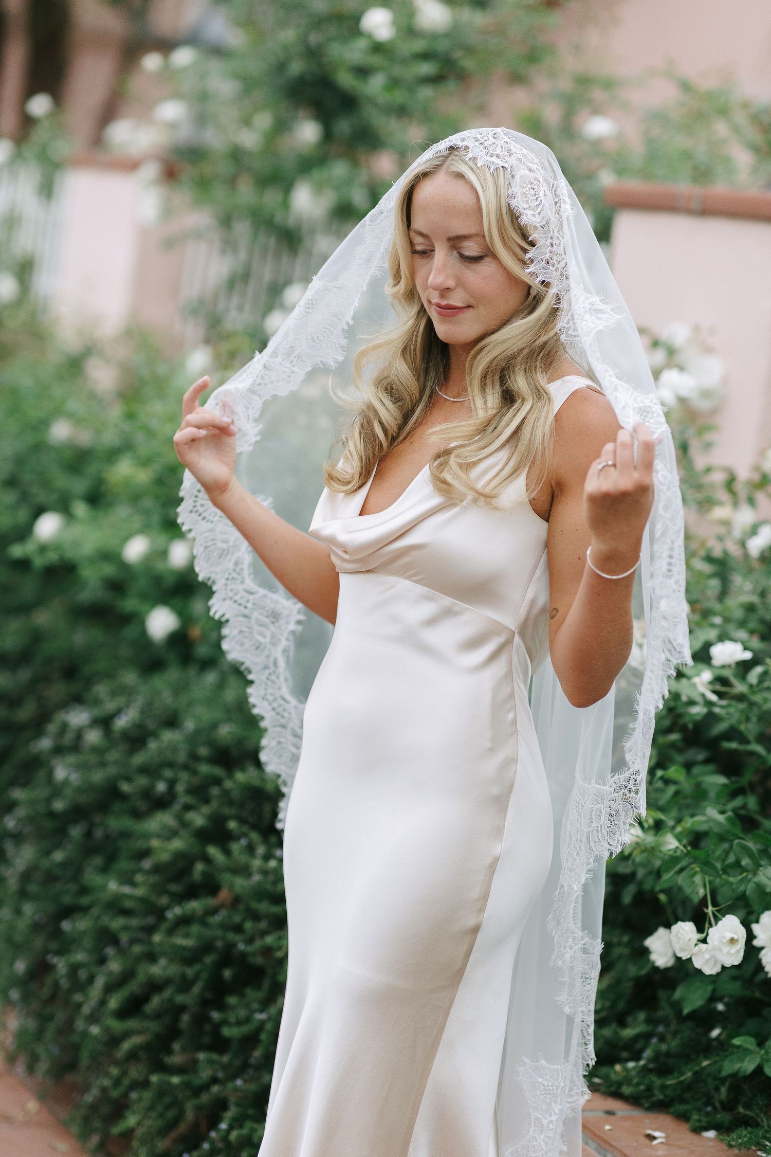 Bride in a fitted white gown with lace veil posing in a garden setting.