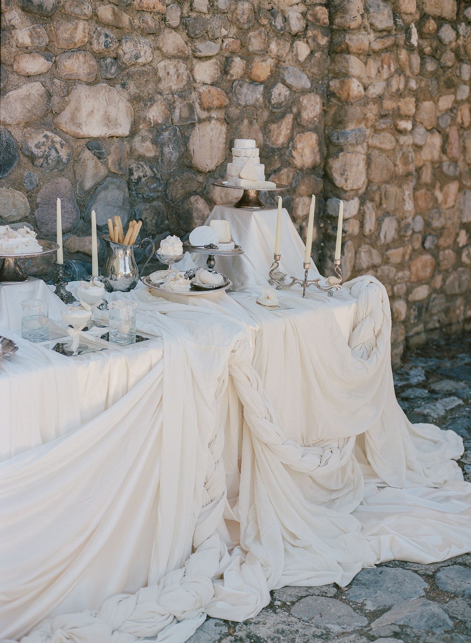 Elegant wedding dessert table draped in ivory fabric with white cake, candles, and silver accents against a stone wall.