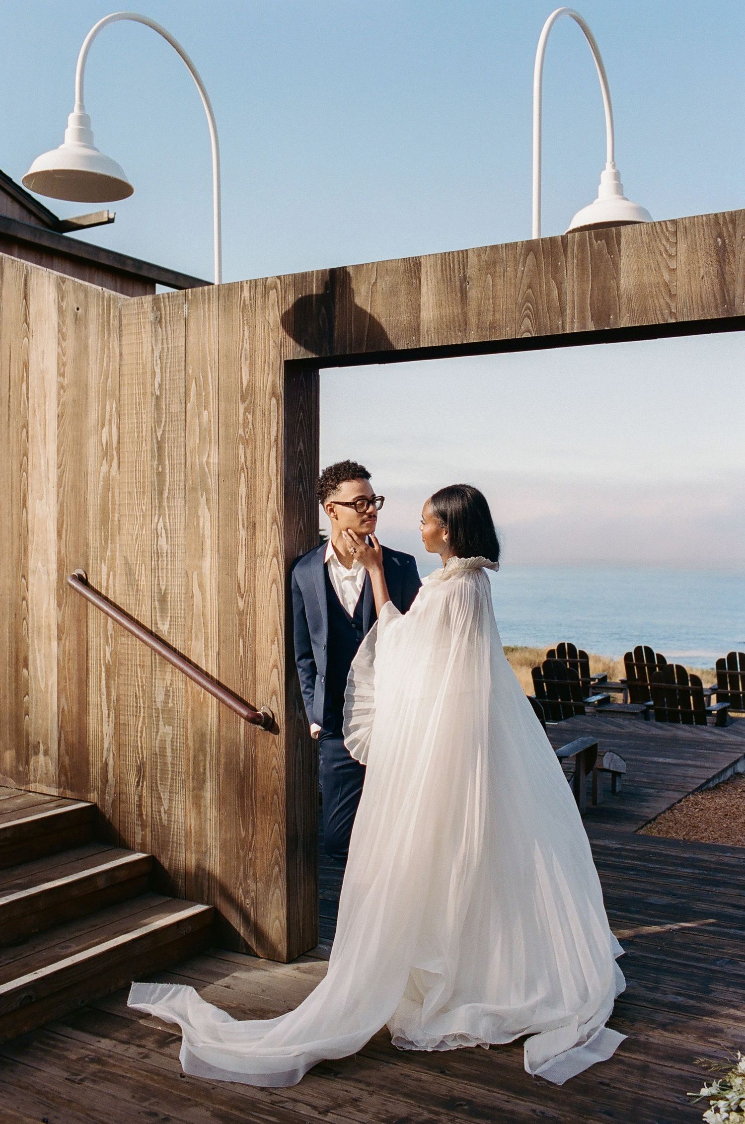 Bride in white cape gown and groom in navy suit share a romantic moment on a wooden deck overlooking the ocean.