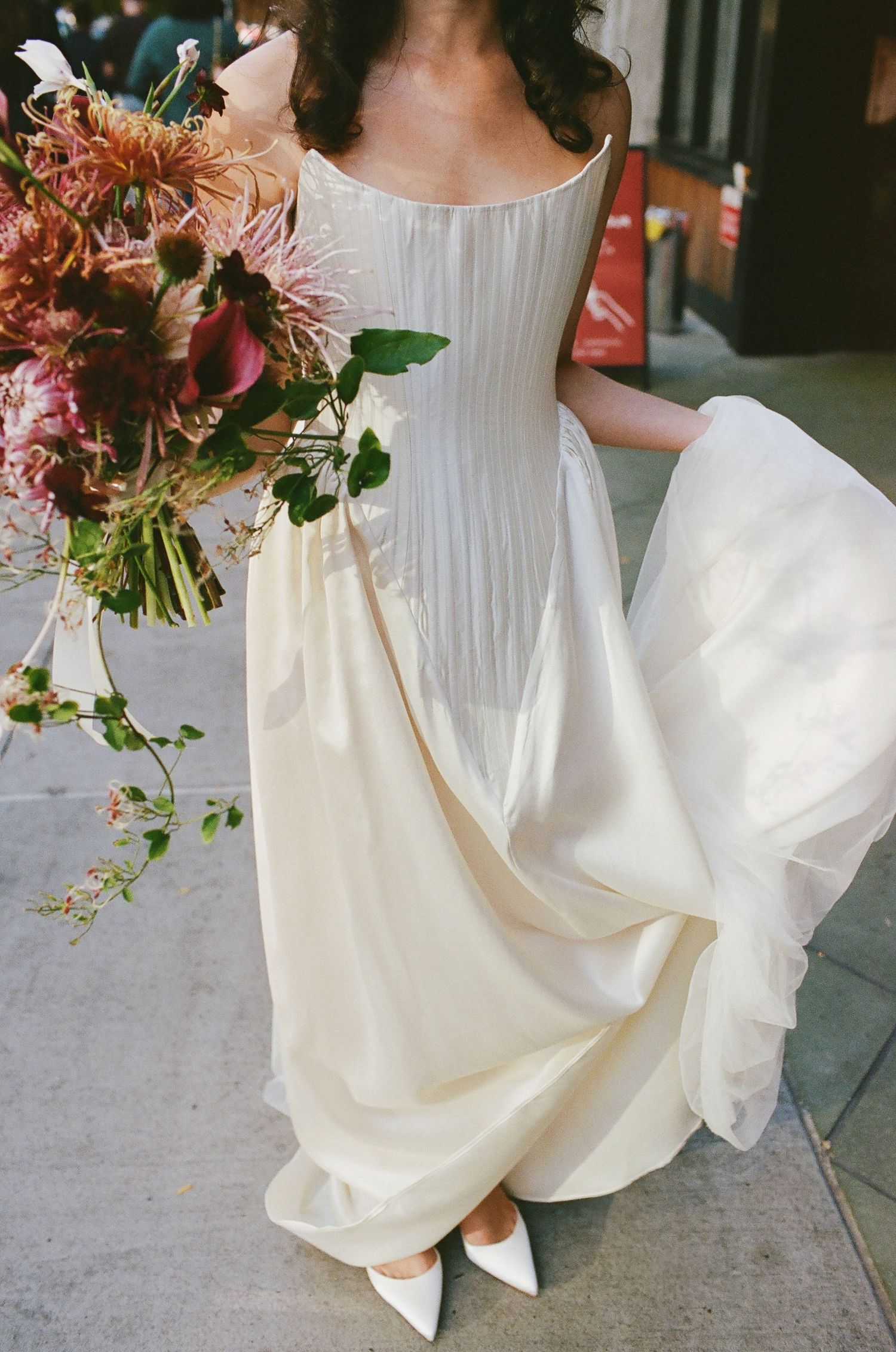 Elegant white pleated wedding dress with flowing skirt and romantic pink and red floral bouquet against urban backdrop.