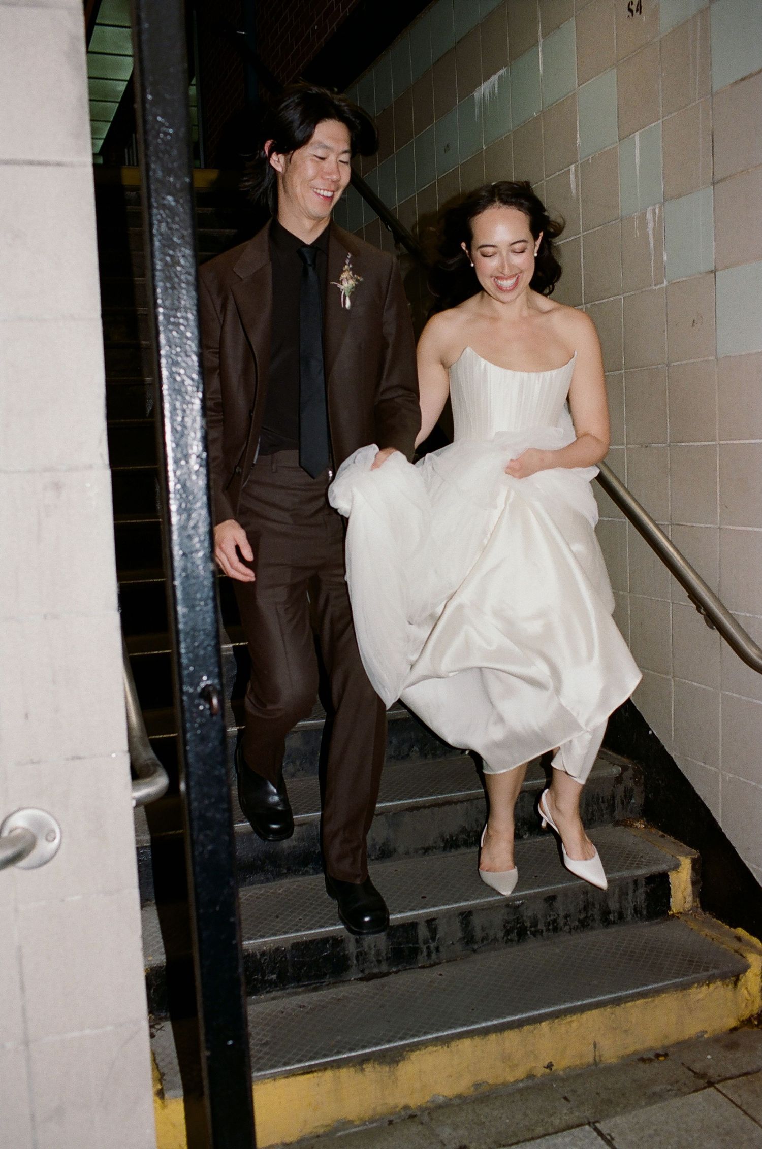 A couple descends concrete stairs together, with her wearing a white strapless gown and him in a brown suit.
