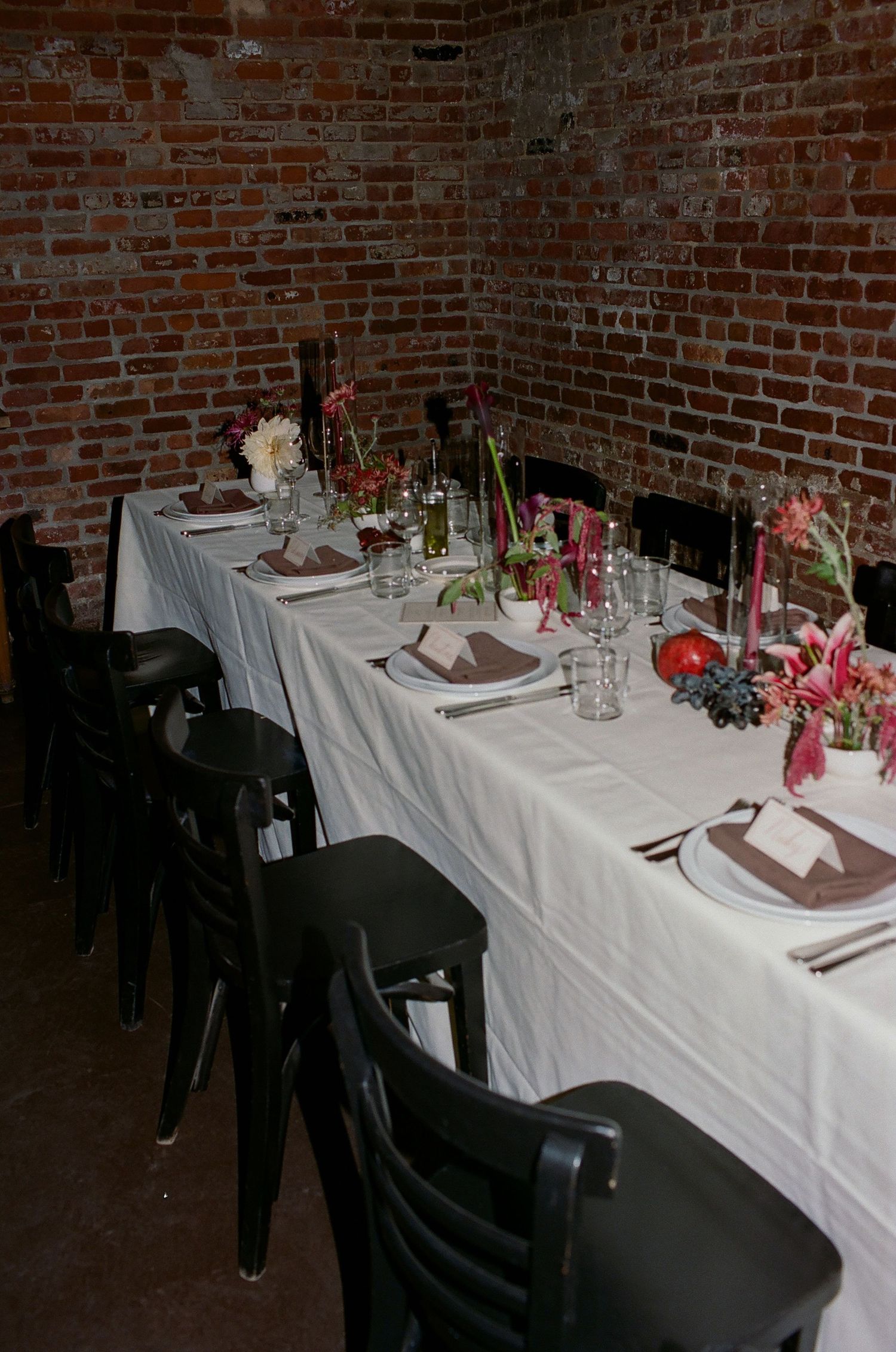 Elegant table setting with white linens, black chairs, and pink floral centerpieces against exposed brick walls in rustic venue.