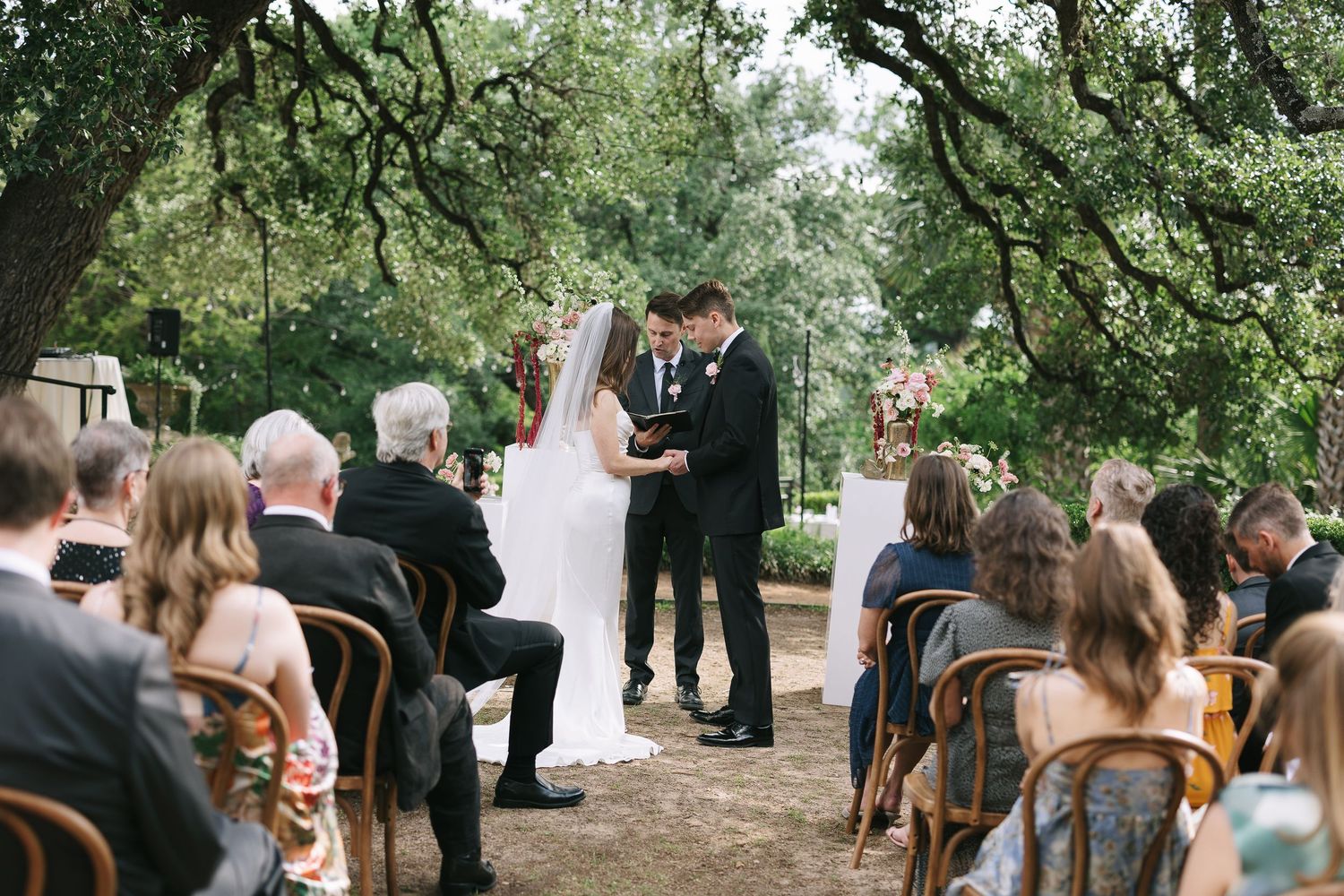 Wedding ceremony processional in a rustic outdoor setting with natural greenery and wooden chairs.