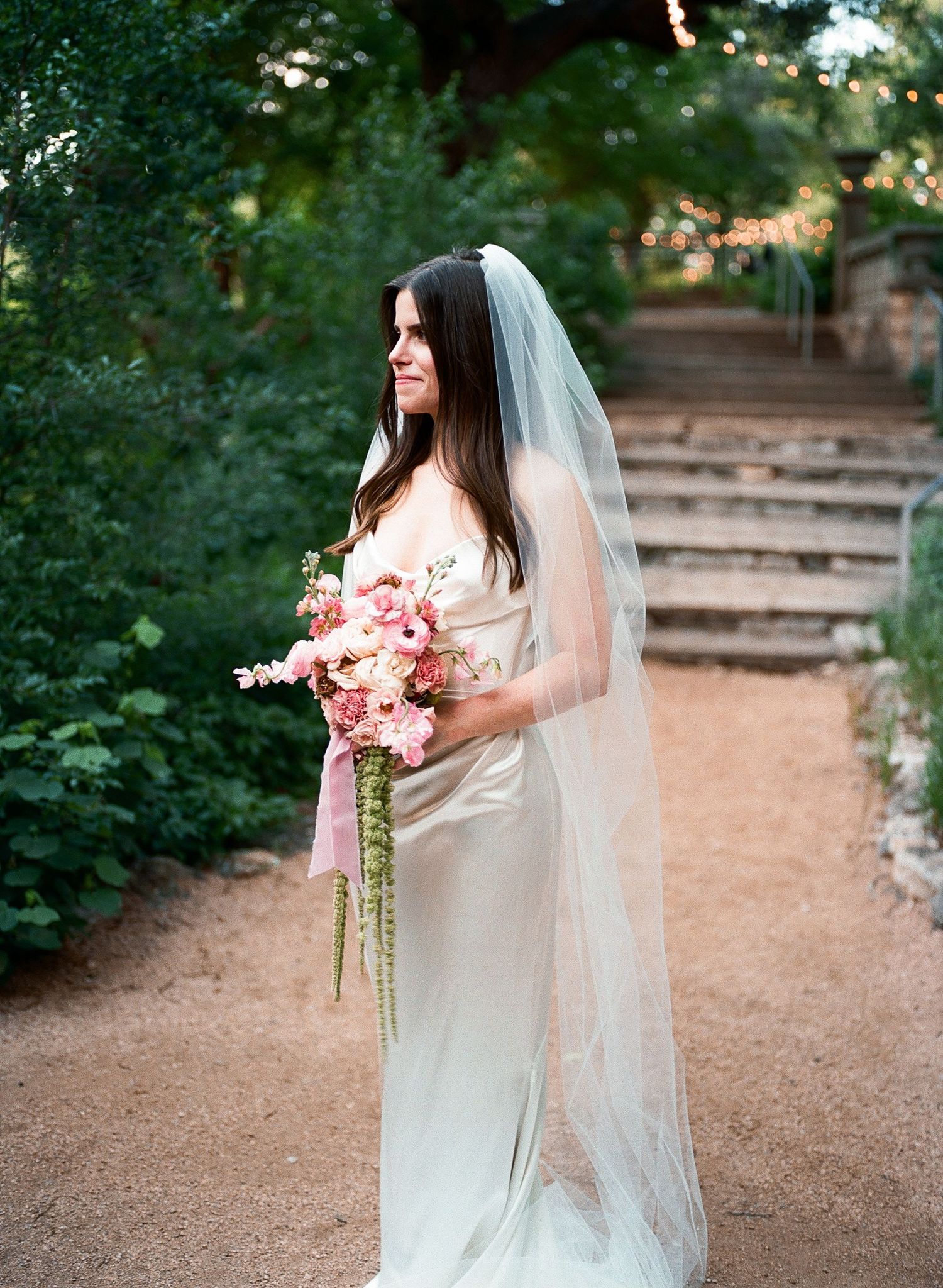 An elegant bridal portrait showcasing a white gown and pink floral bouquet in an outdoor setting.