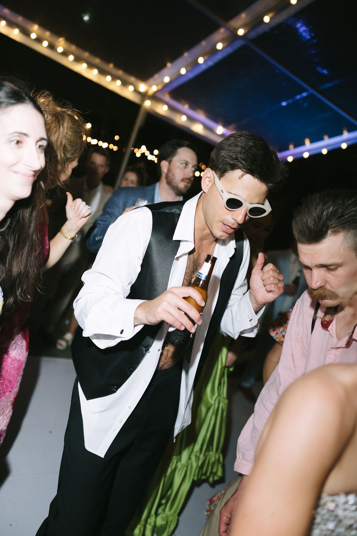 Partygoers dancing and socializing at a nighttime event under string lights with someone in sunglasses and formal attire.