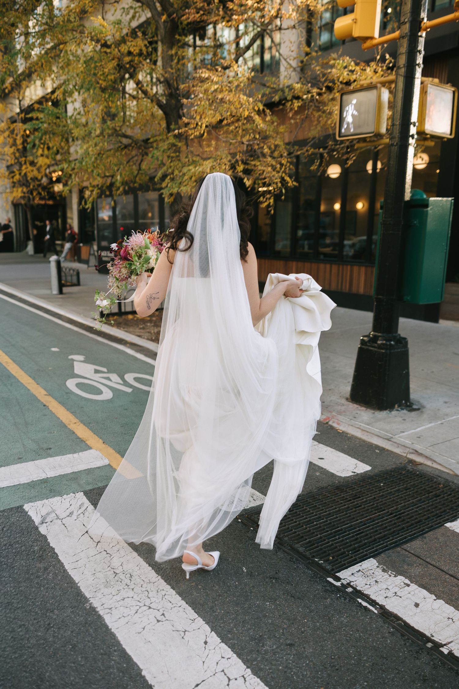 A flowing white wedding dress and veil photographed in motion on a city sidewalk with autumn trees in the background.