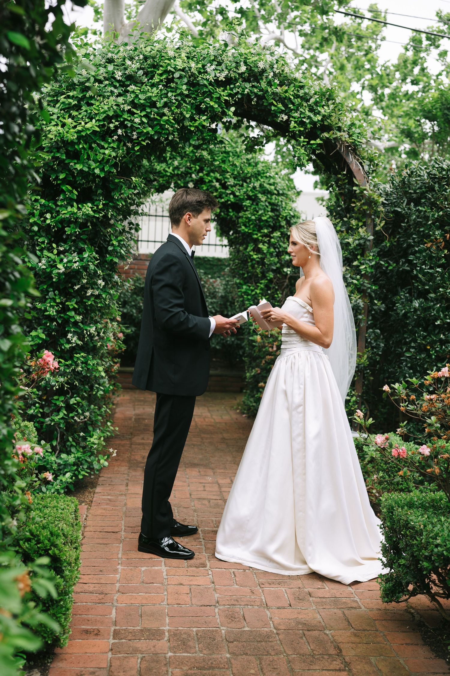 A wedding couple shares a moment on a brick pathway surrounded by green hedges and roses.