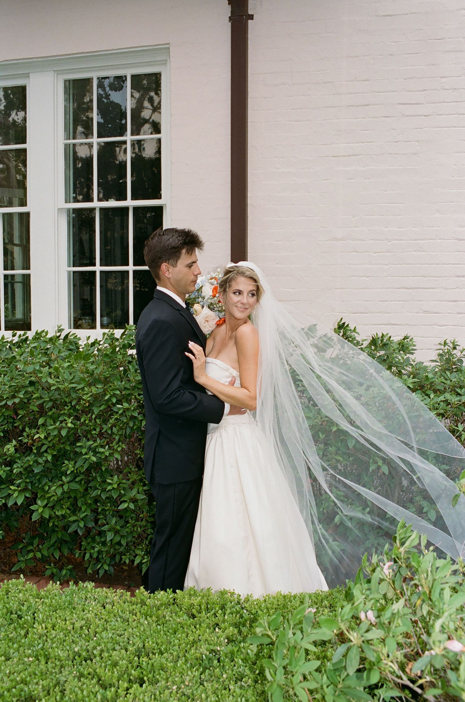 A couple in wedding attire stands together as the bride's veil flows in the breeze.