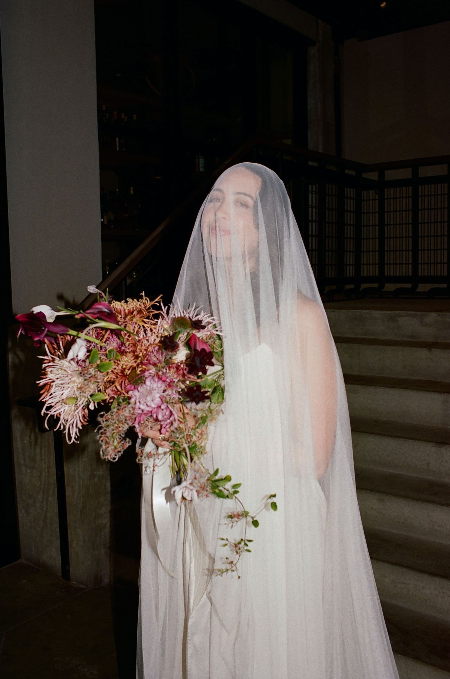 A bride in a flowing white veil holds a vibrant bouquet of pink and burgundy flowers on a staircase.