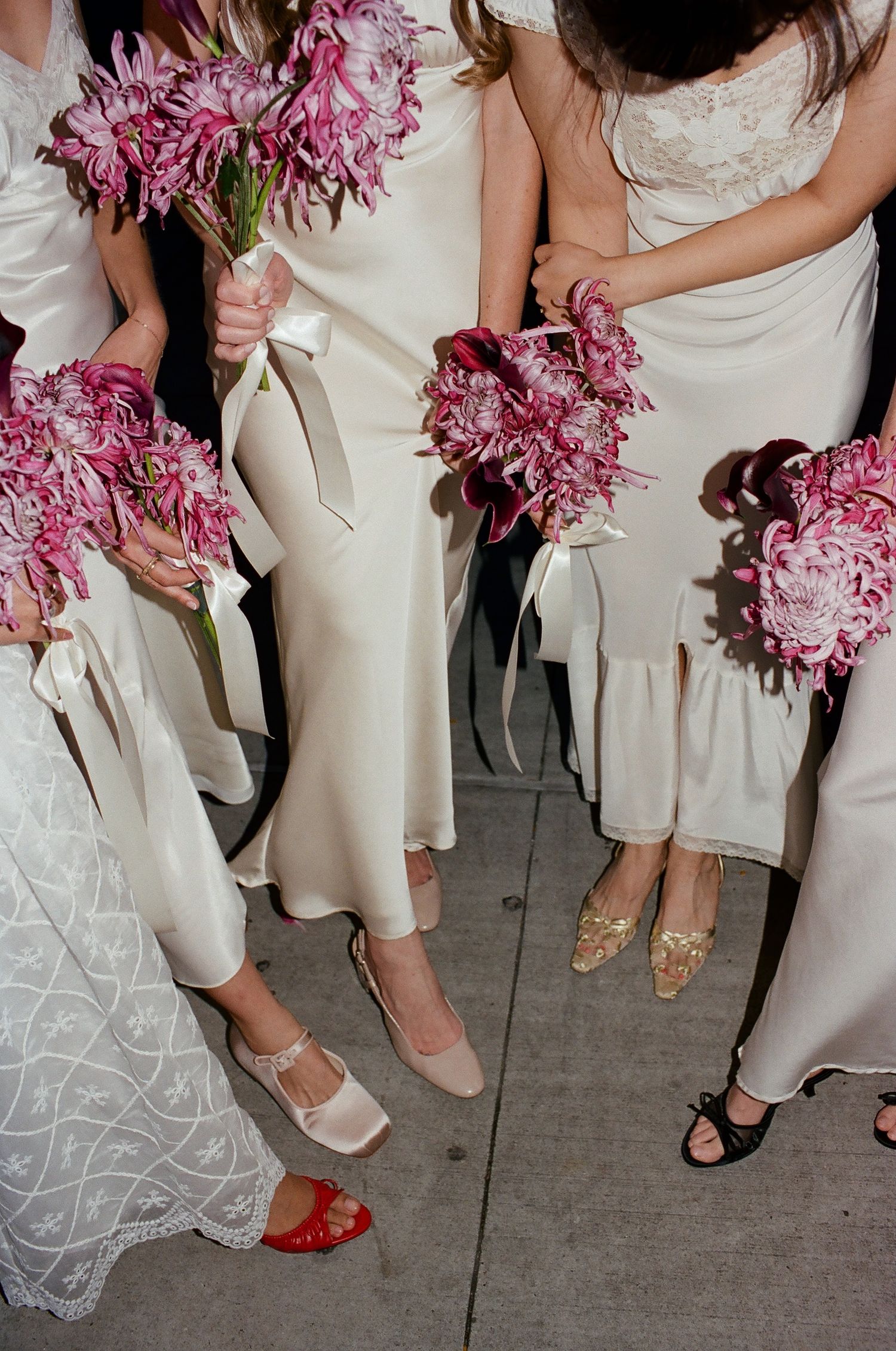 Bridesmaids in white satin dresses holding pink chrysanthemum bouquets with ribbon, wearing varied heels on pavement.