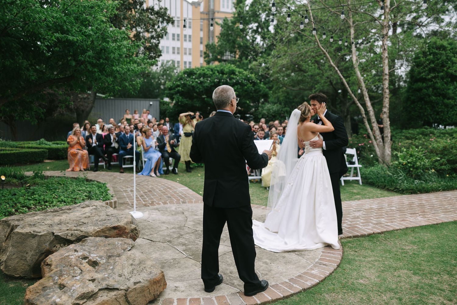 Wedding party walking along a curved brick pathway in a garden setting during a celebration.