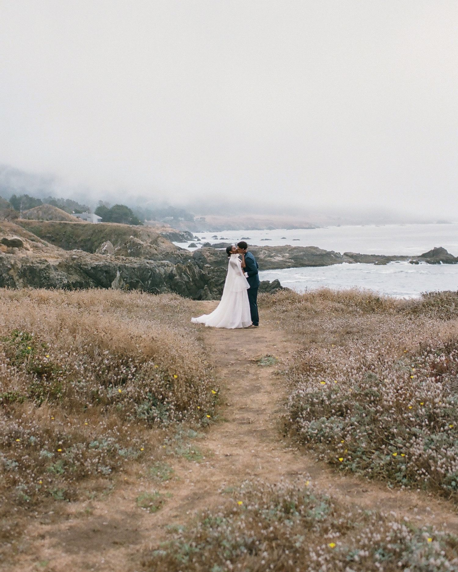 Bride and groom embrace on a coastal trail surrounded by golden wildflowers and misty ocean views on their wedding day.