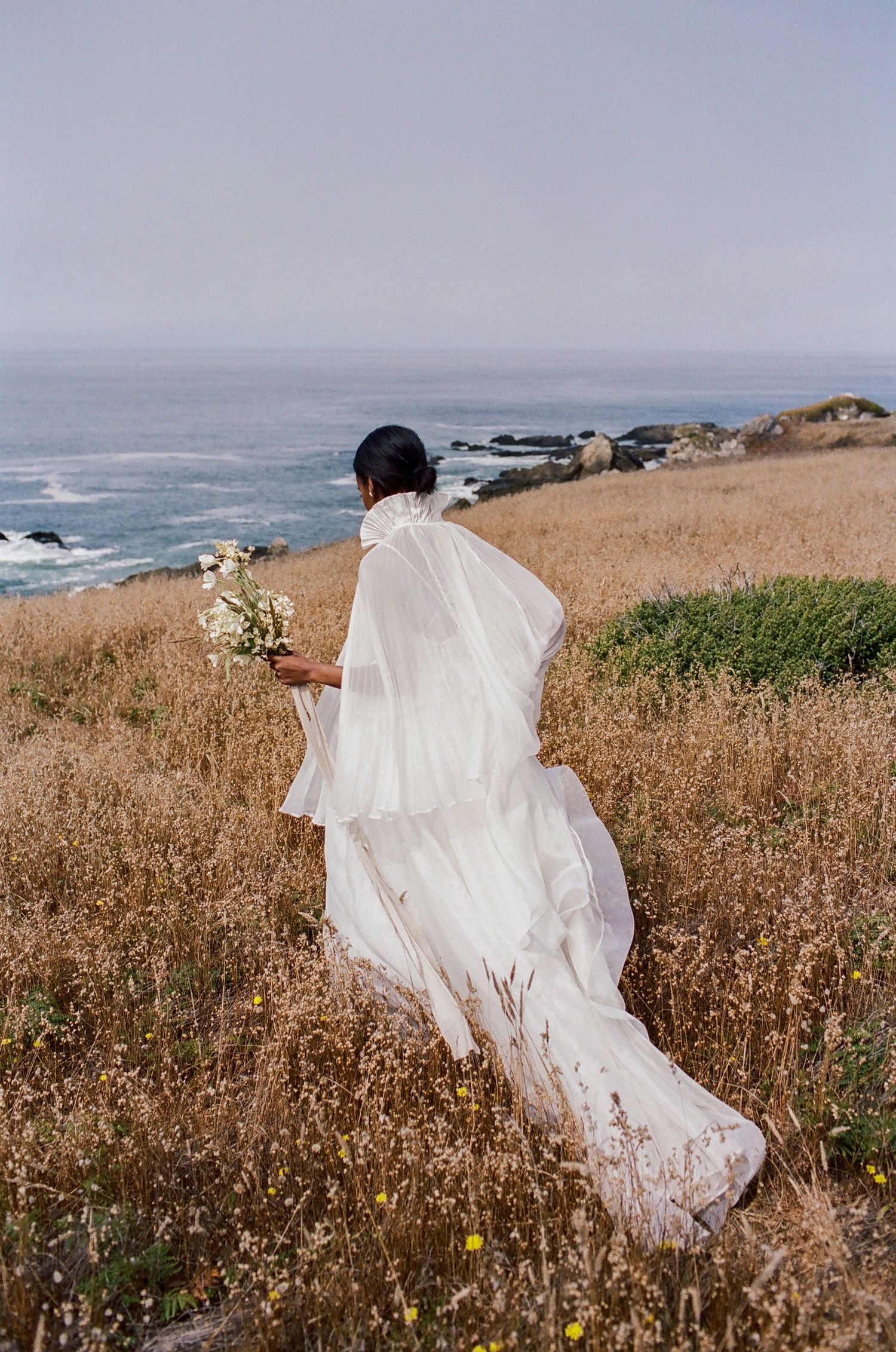 Bride in flowing white gown and veil walks through golden coastal meadow holding bouquet, ocean vista behind her.