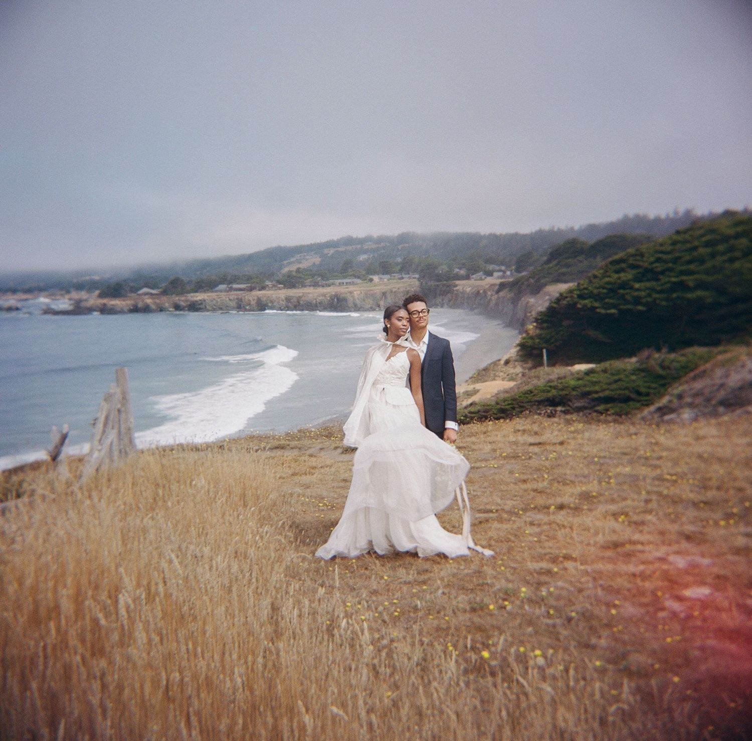 Bride and groom posing on a coastal bluff with ocean waves and foggy shoreline in the background.