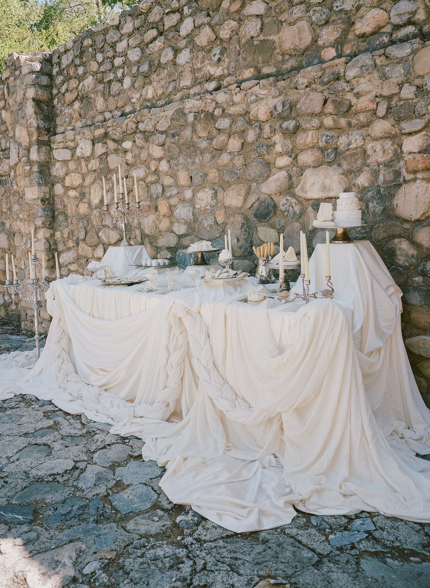 Elegant outdoor wedding dessert table draped in ivory fabric against a rustic stone wall with candles and decorative accents.