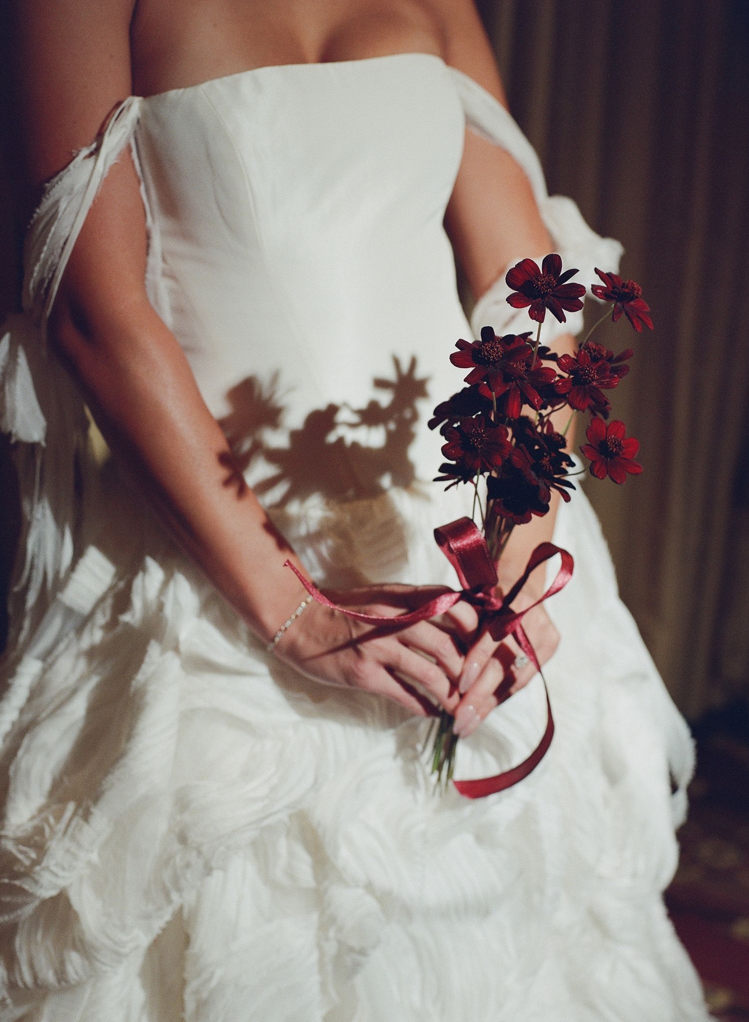 Bride in white strapless gown holding small red floral bouquet tied with satin ribbon, flower shadows on dress.