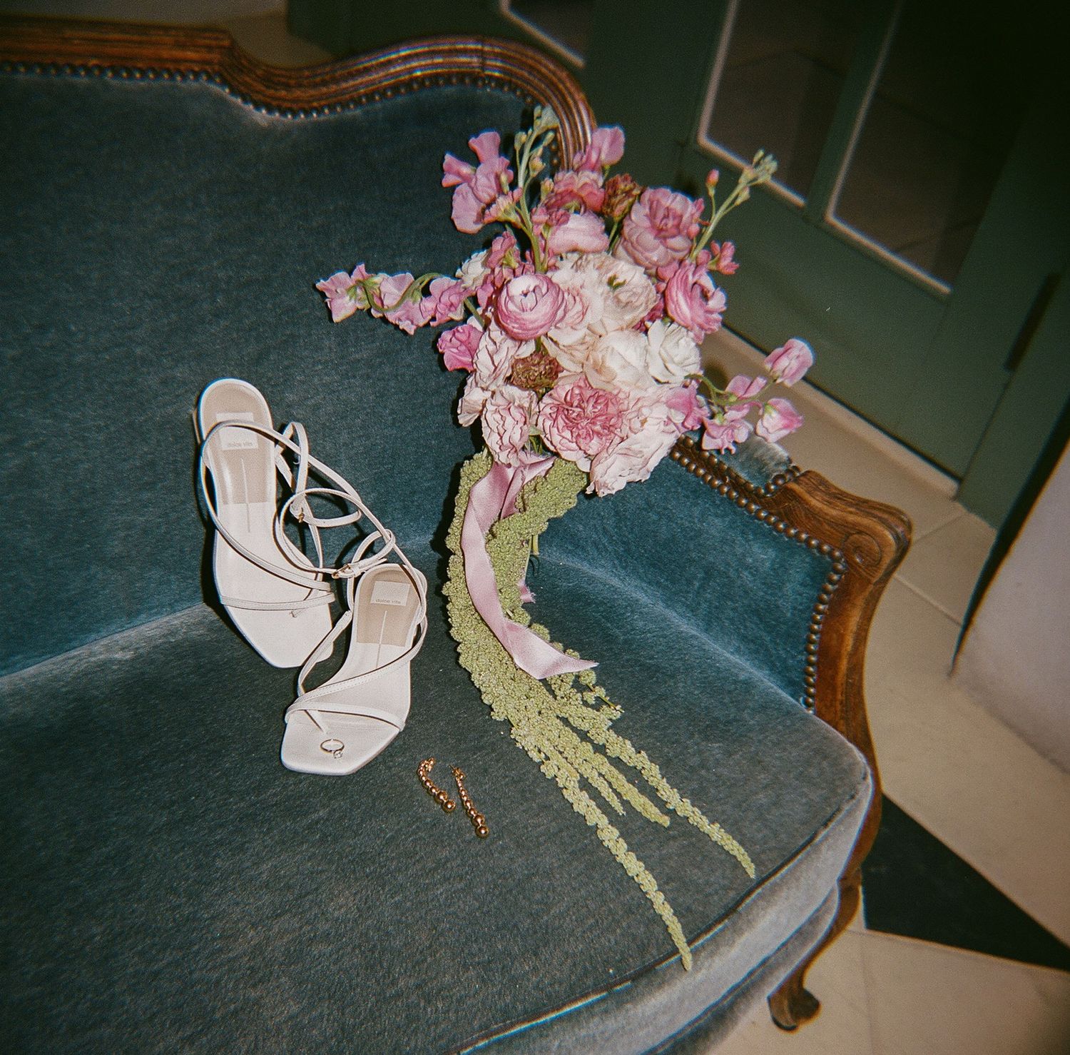 White strappy wedding shoes next to a pink floral bouquet on blue velvet furniture.