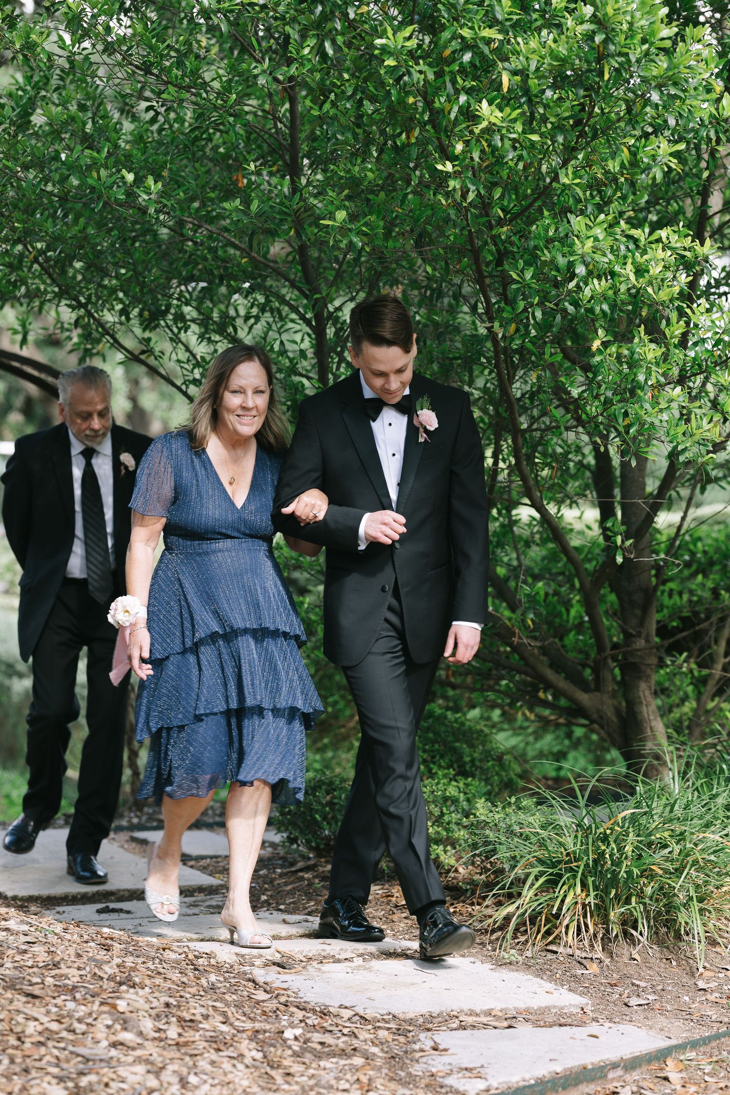 Wedding guests in formal attire walk along a garden path during an outdoor ceremony.