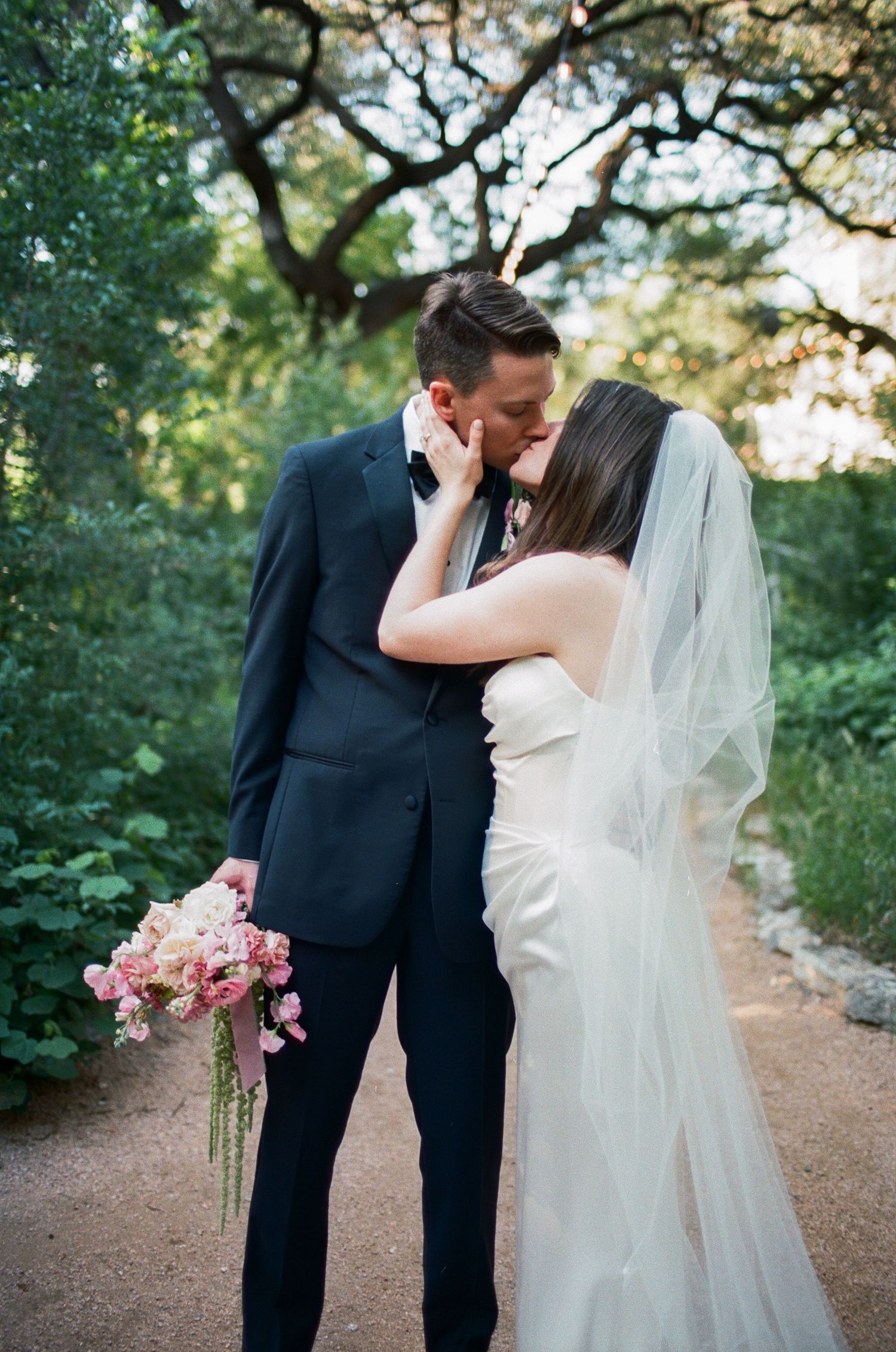 A romantic wedding couple shares intimate moments on a tree-lined path with soft natural lighting.