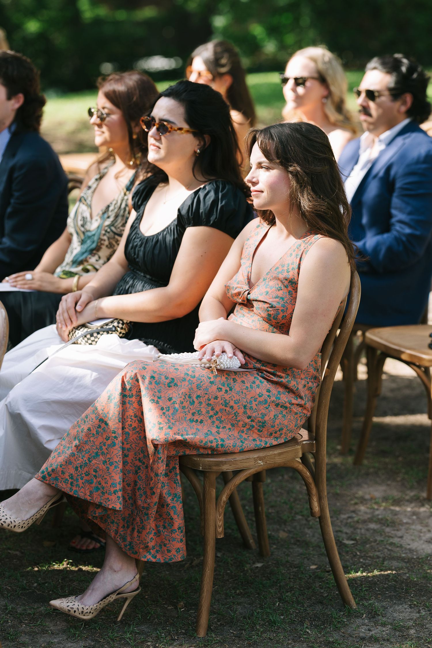 Wedding guests in floral dresses seated on wooden chairs at outdoor ceremony.