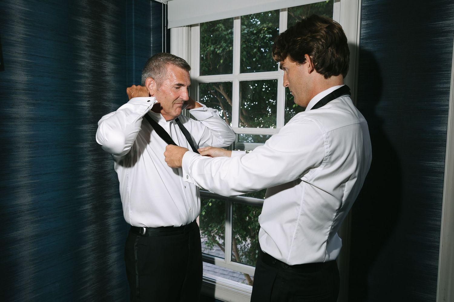 Two people in formal wear interact playfully in a dark room with white trim windows.