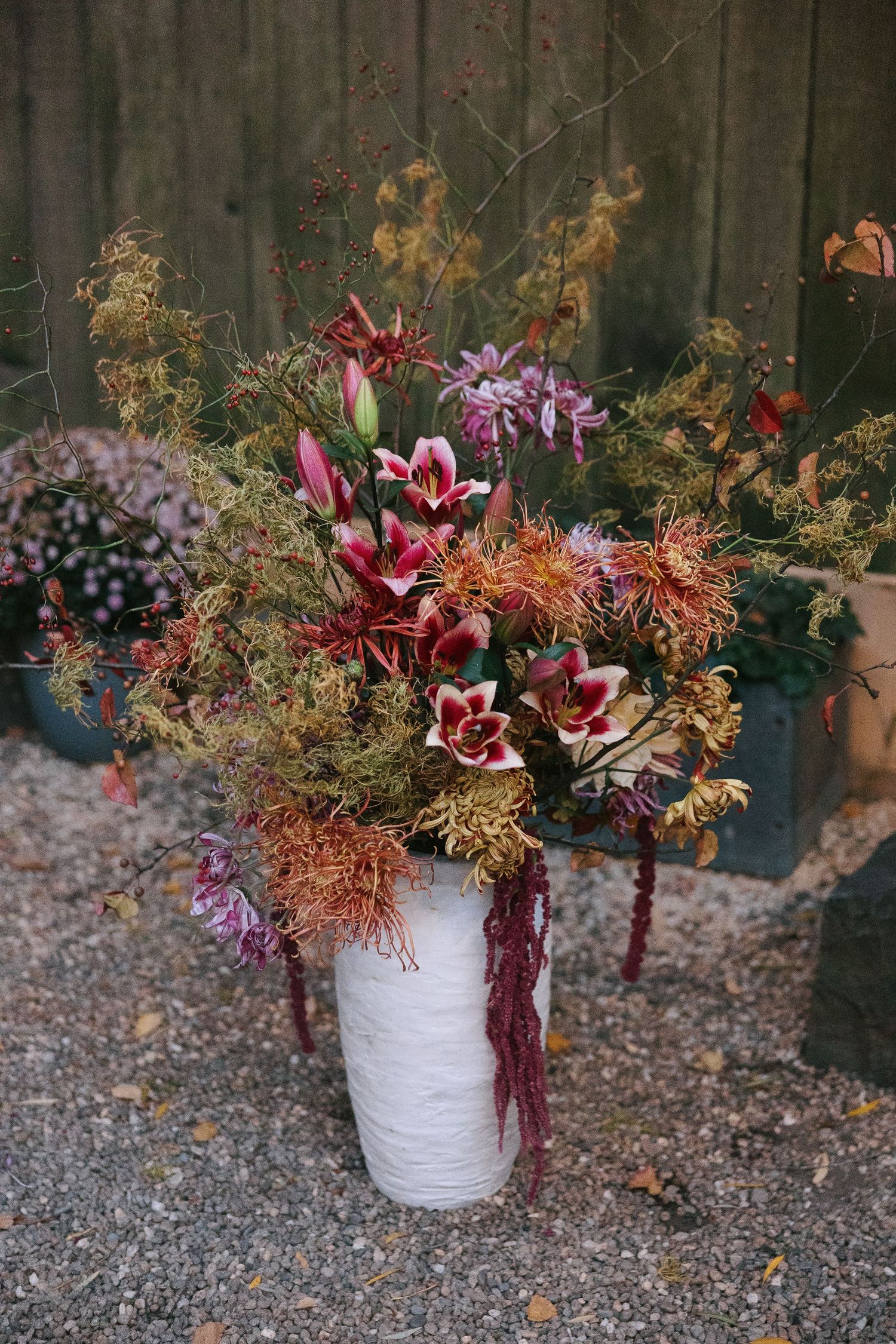 Large floral arrangement in white vase featuring lilies and autumn foliage against wooden fence.