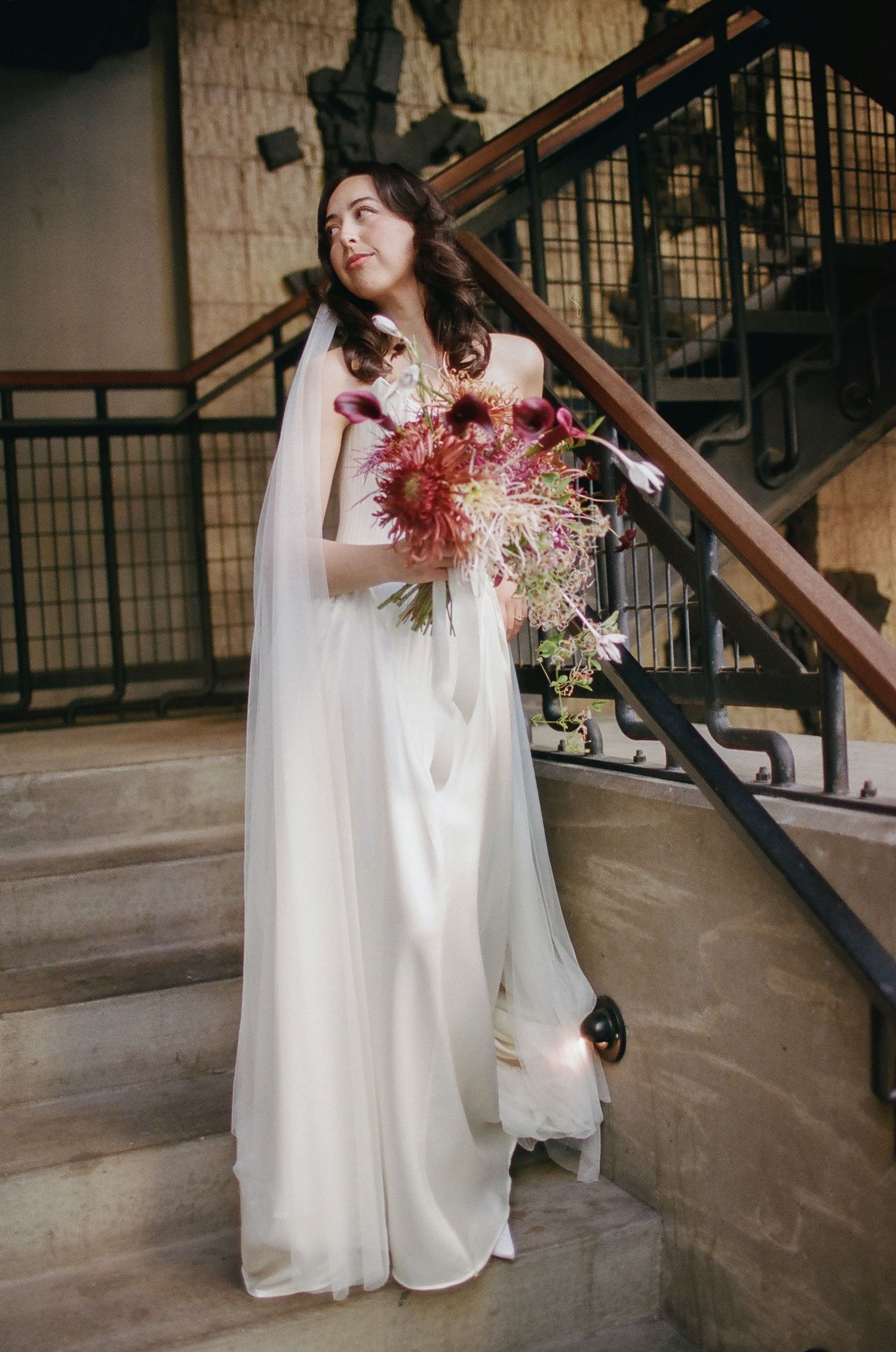 A bride in a flowing white dress and cape holds a vibrant pink bouquet while standing near an industrial staircase.