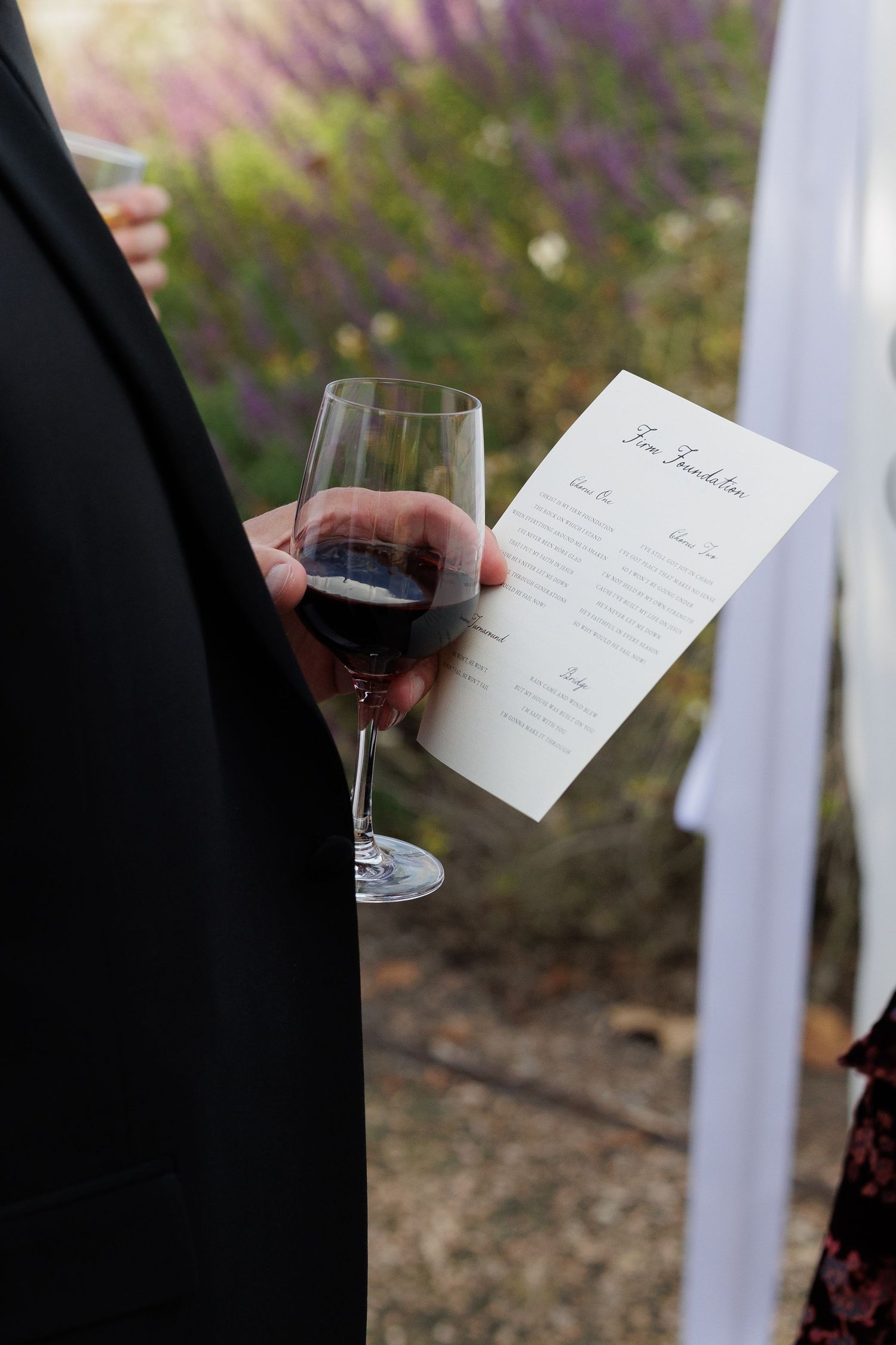 Person in black suit holding red wine glass and wedding ceremony program outdoors near lavender bushes.