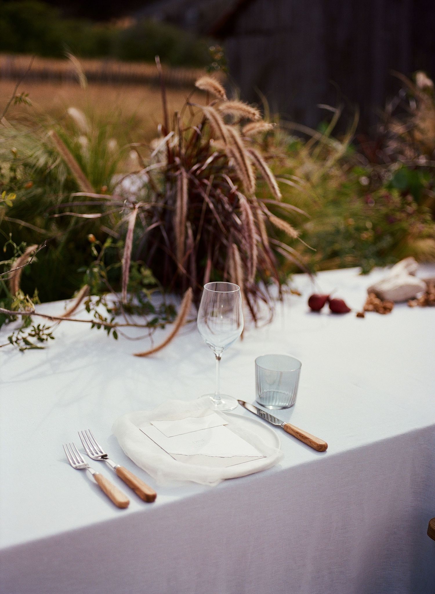 Elegant outdoor place setting with white linen, wine glass, wooden cutlery, and wild grass centerpiece.